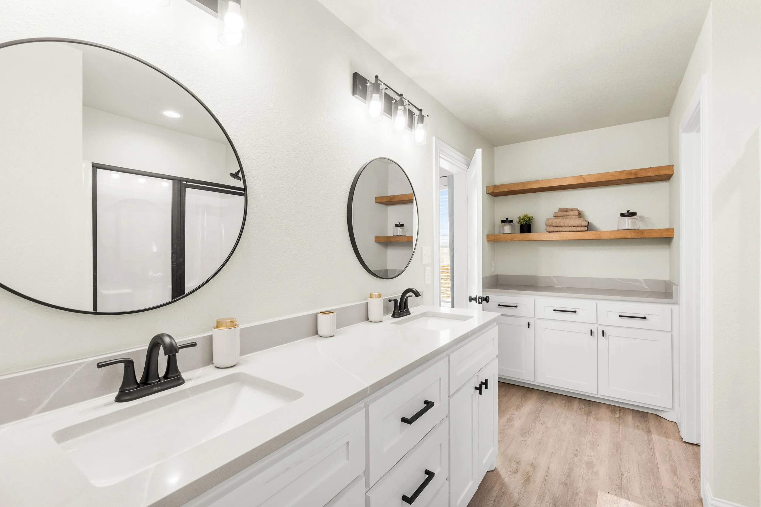 Modern bathroom with double vanity, large round mirrors, black fixtures, white cabinets, wooden open shelves with towels and jars, and a closet with a glass sliding door.