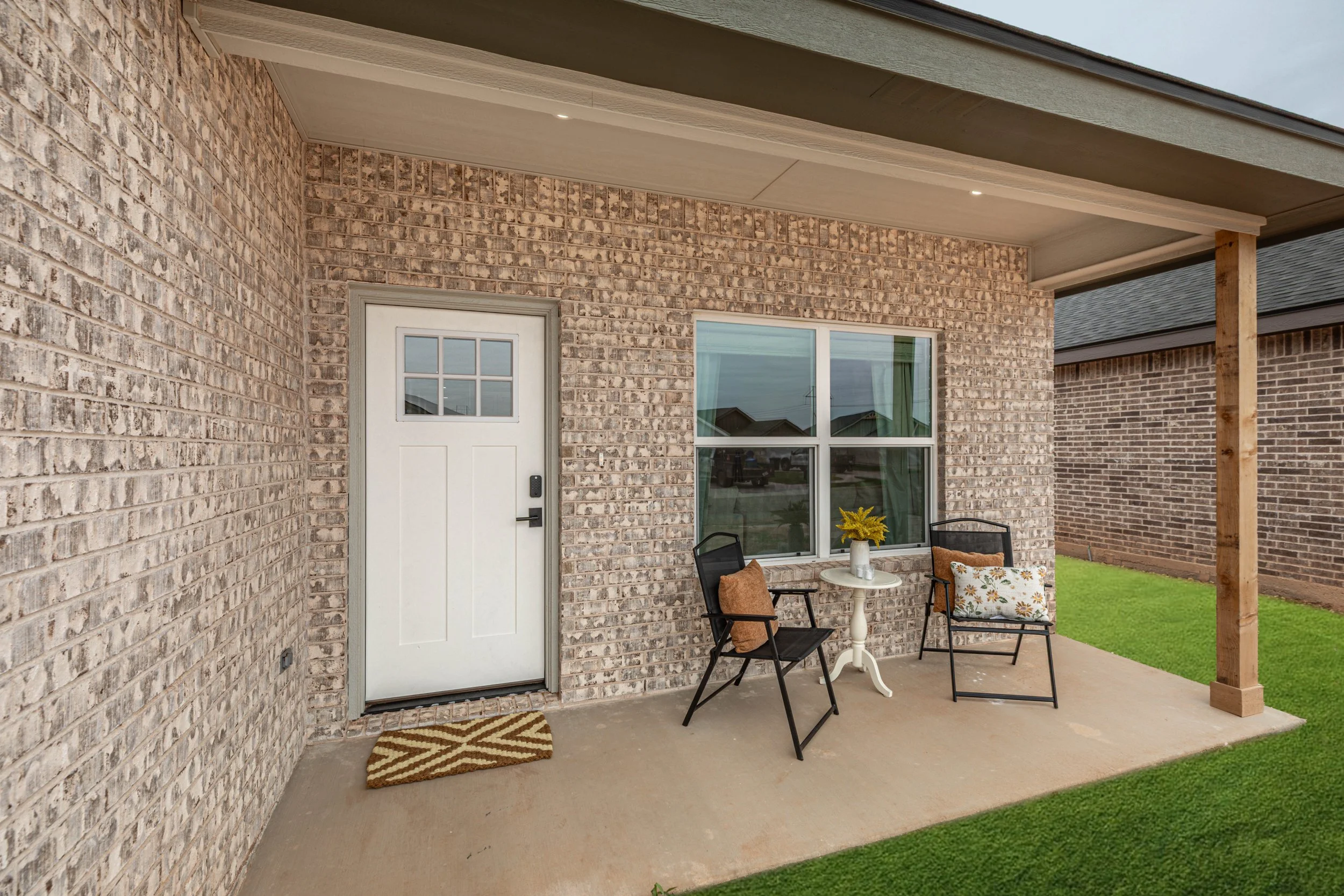 Back porch of a brick house with a white door, window, small white table with flowers, and two chairs.