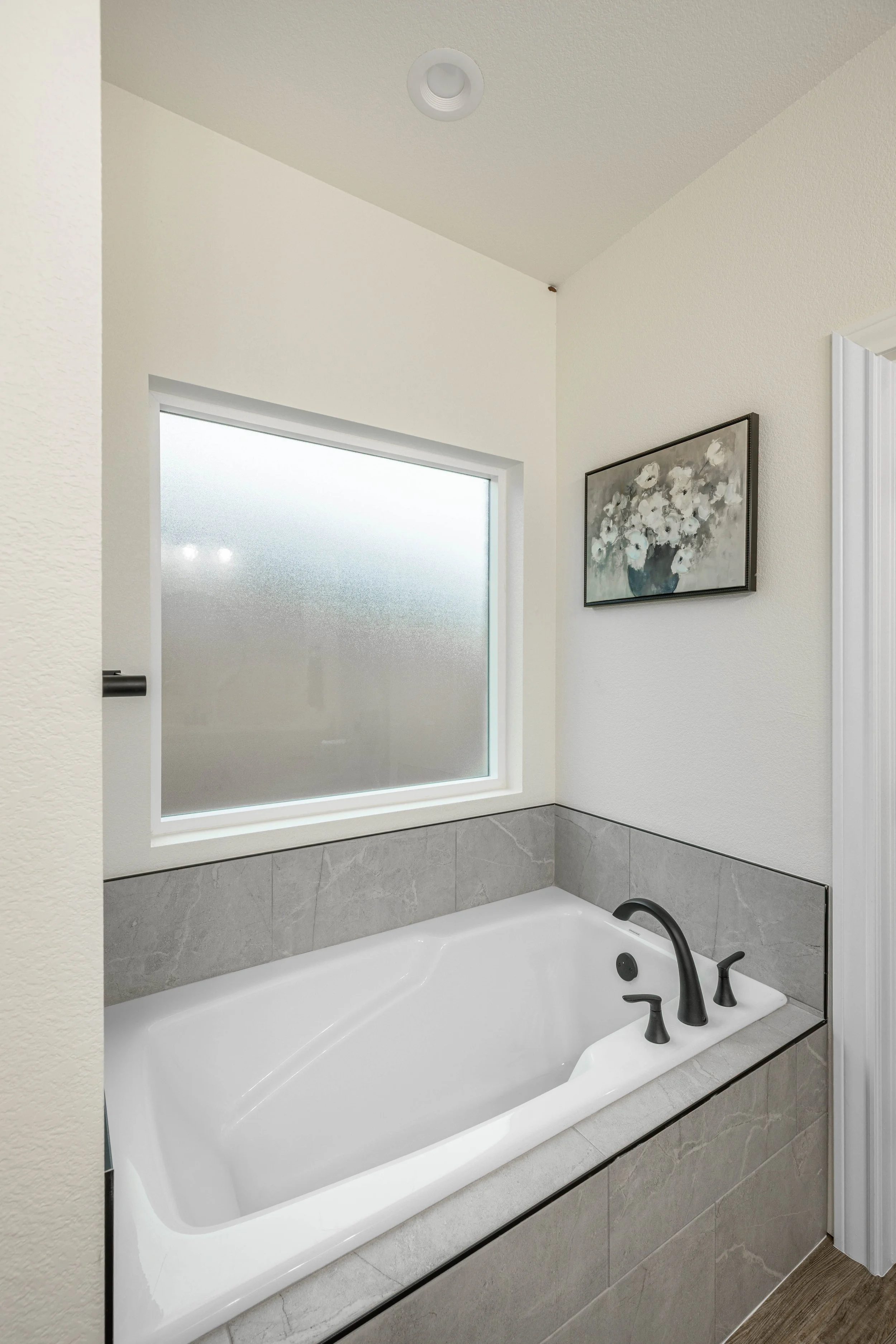 Bathroom with bathtub, gray tile surround, black faucet, frosted glass window, floral artwork on the wall, white walls, ceiling light fixture.