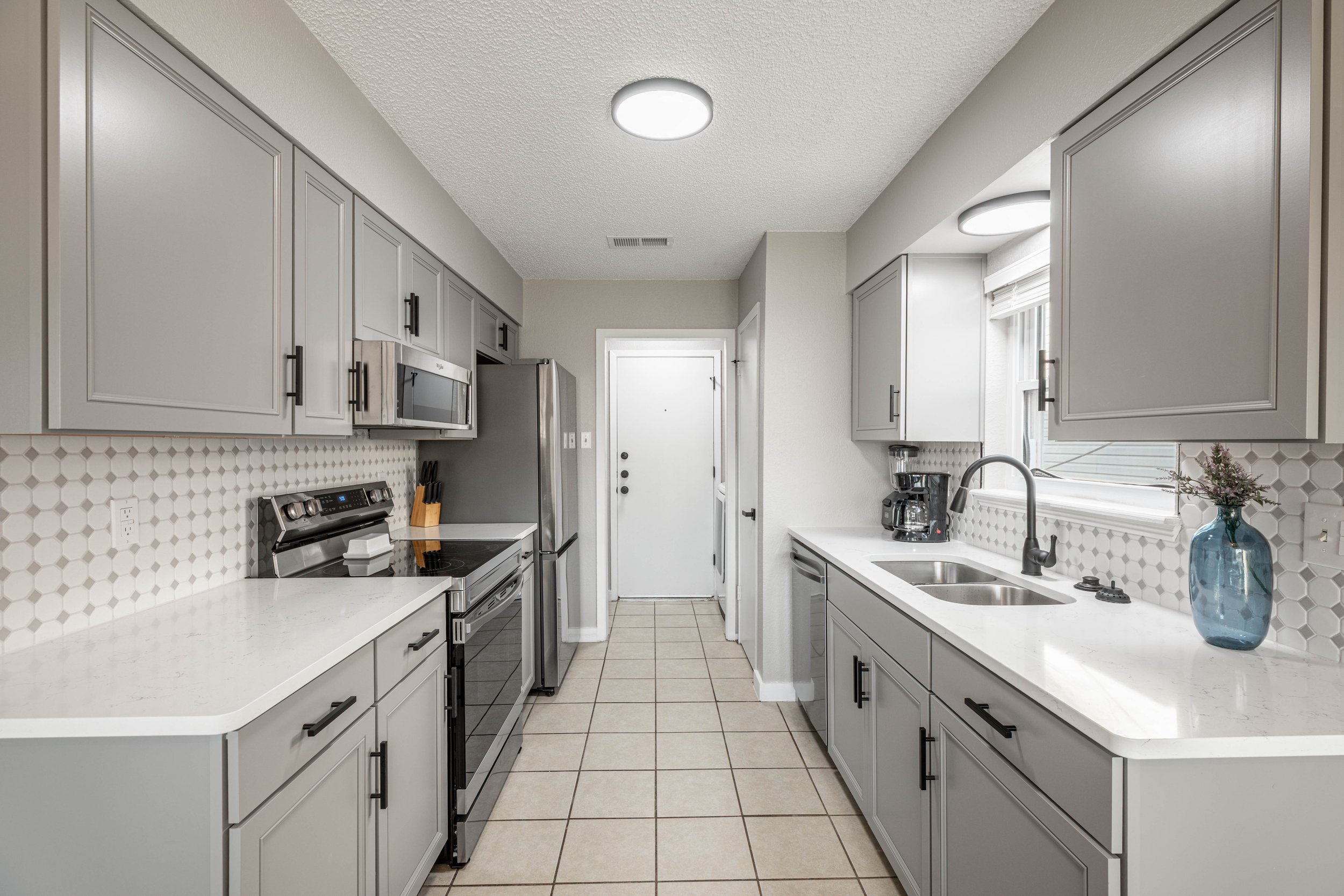 A modern kitchen with white countertops, gray cabinets, stainless steel appliances, a tiled backsplash, and a window with a blind. There is a blue vase with flowers on the counter.