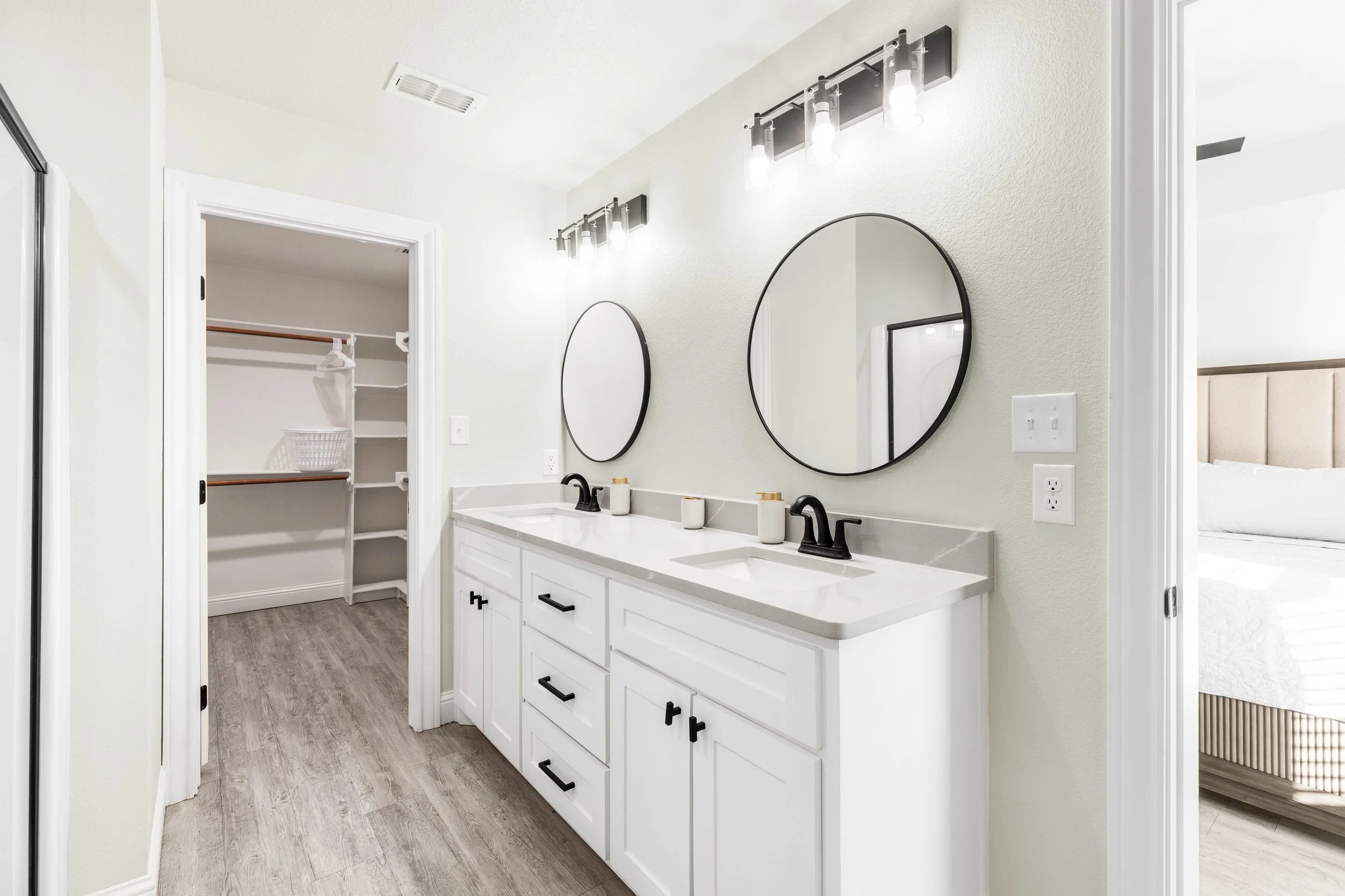 A modern bathroom with a double vanity, rectangular sinks, black fixtures, and oval mirrors, with a walk-in closet visible in the background.