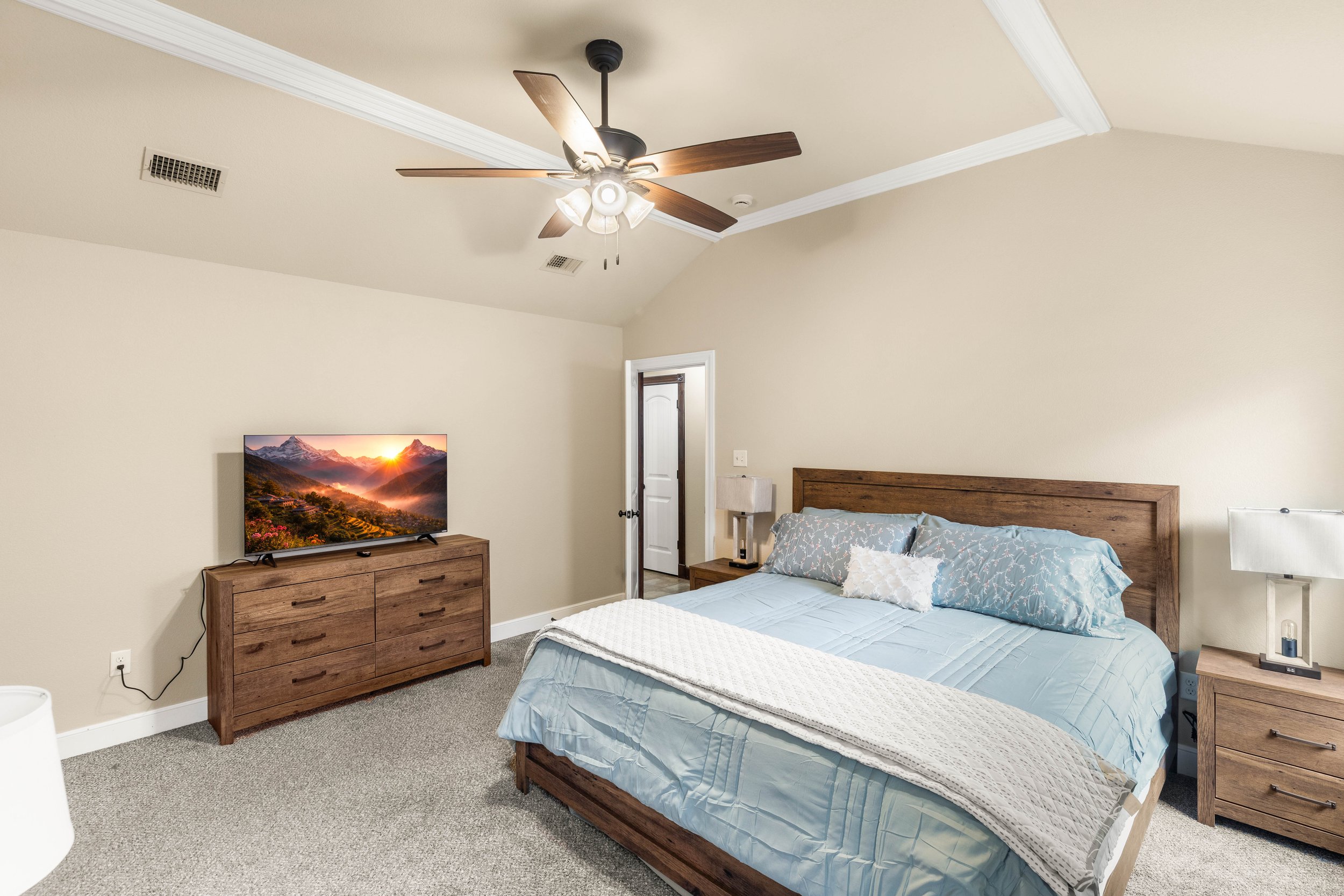 A bedroom with a wooden bed, light blue bedding, matching pillows, a wooden dresser with a television, and a nightstand with a lamp. The ceiling has a fan with wooden blades.