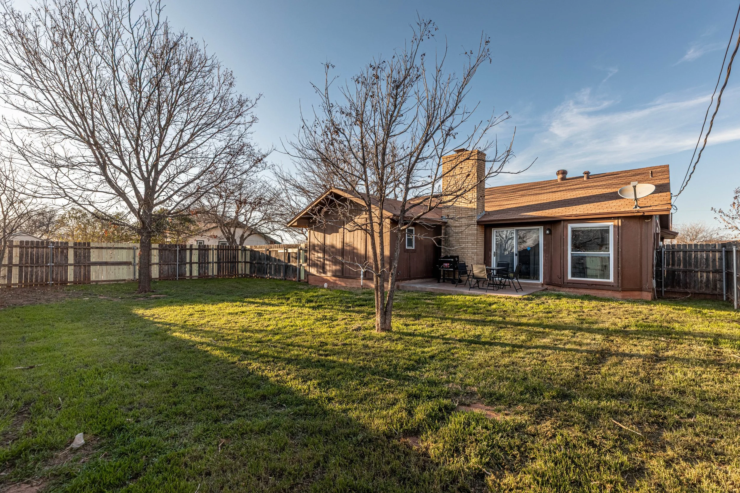 A backyard scene with a grassy lawn, a wooden fence, leafless trees, and a house with a brick chimney, sliding glass door, and patio furniture under a blue sky with some clouds.