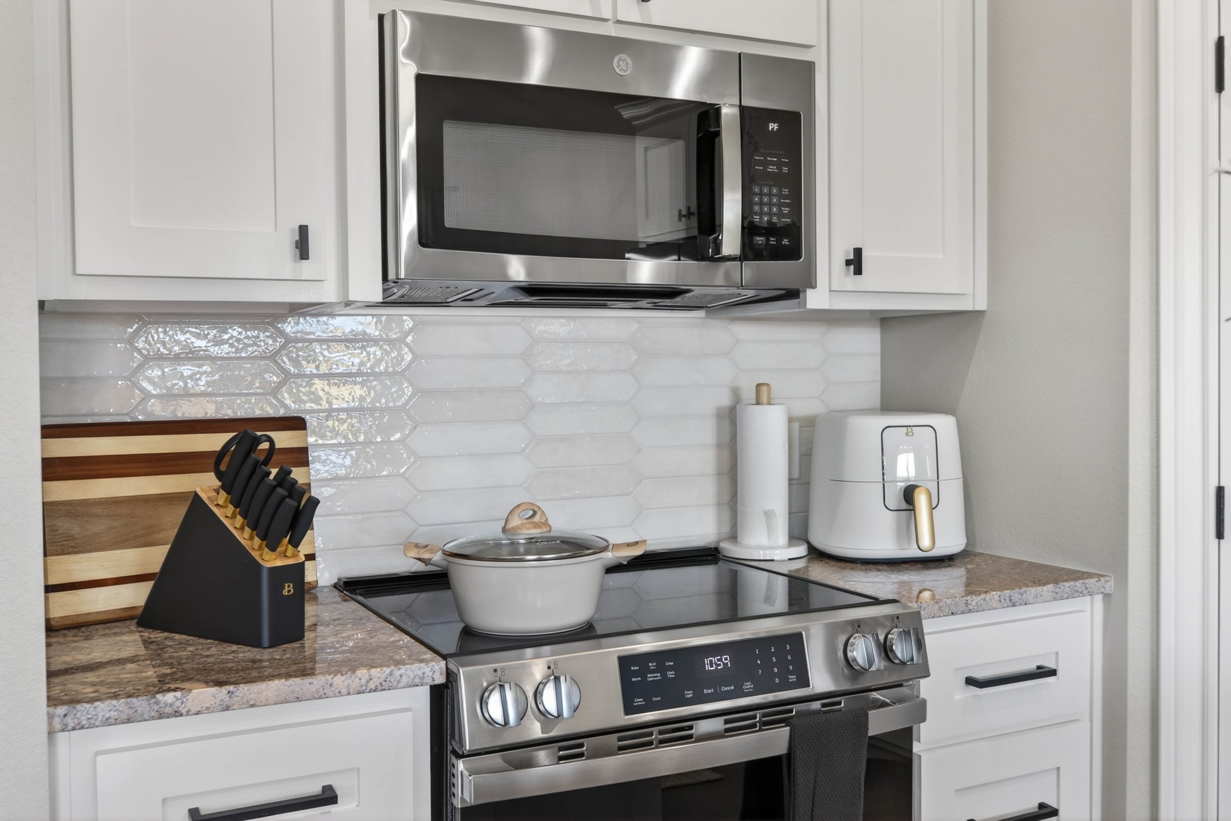 A modern kitchen with stainless steel microwave above a stove, white cabinets, a white toaster, a paper towel roll, a knife block, and a white pot on the stove.