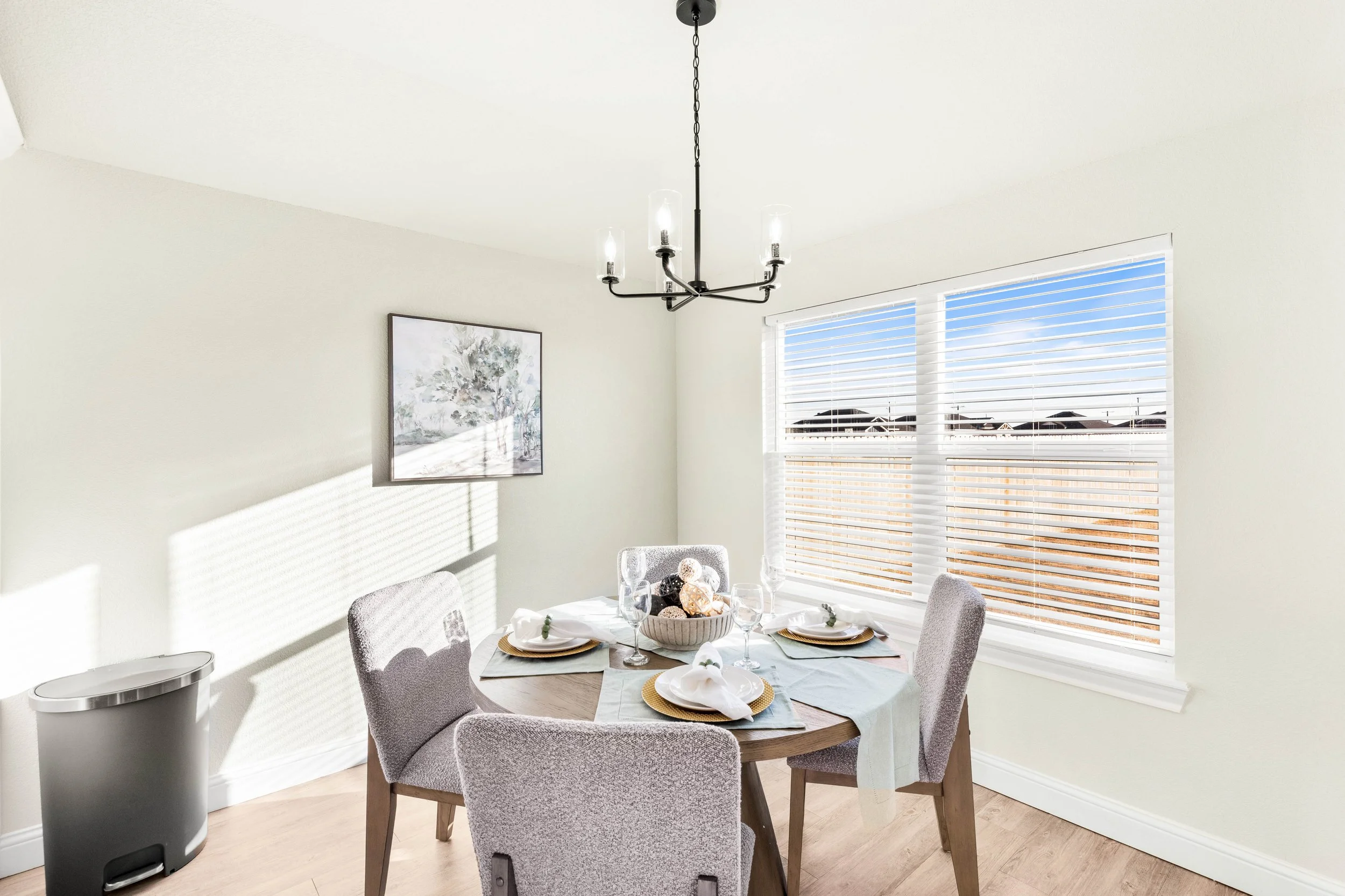 A dining room with a round table set with white dishes, glasses, and napkins, surrounded by four upholstered chairs. There is a modern black chandelier hanging from the ceiling and a window with blinds letting in sunlight, casting shadows on the wall