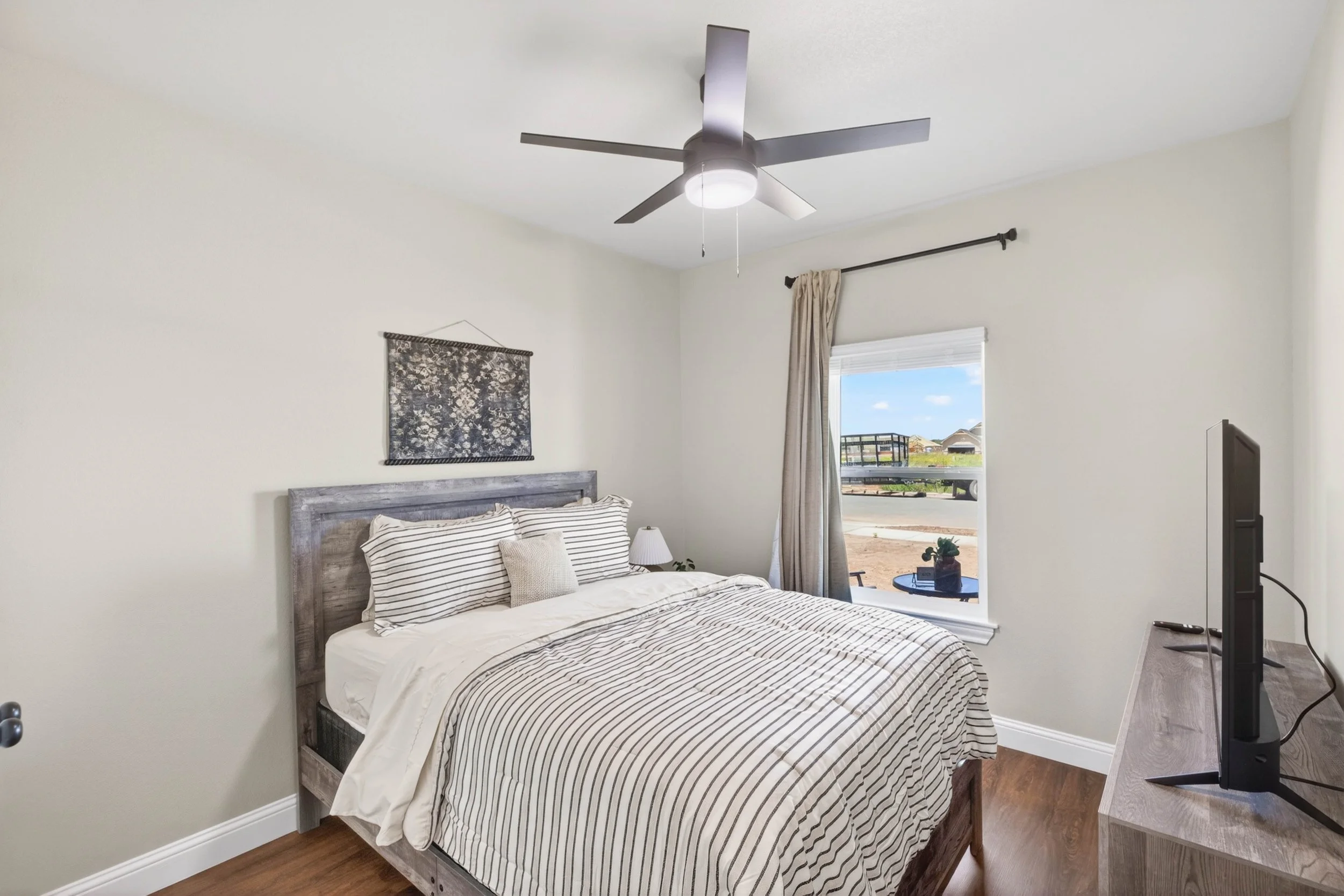 A bedroom with a wooden bed frame, striped bedding, a small bedside table with a lamp, a picture hanging on the wall, a window with beige curtains, and a view outside showing a trailer and houses. A ceiling fan with lights hangs from the white ceilin