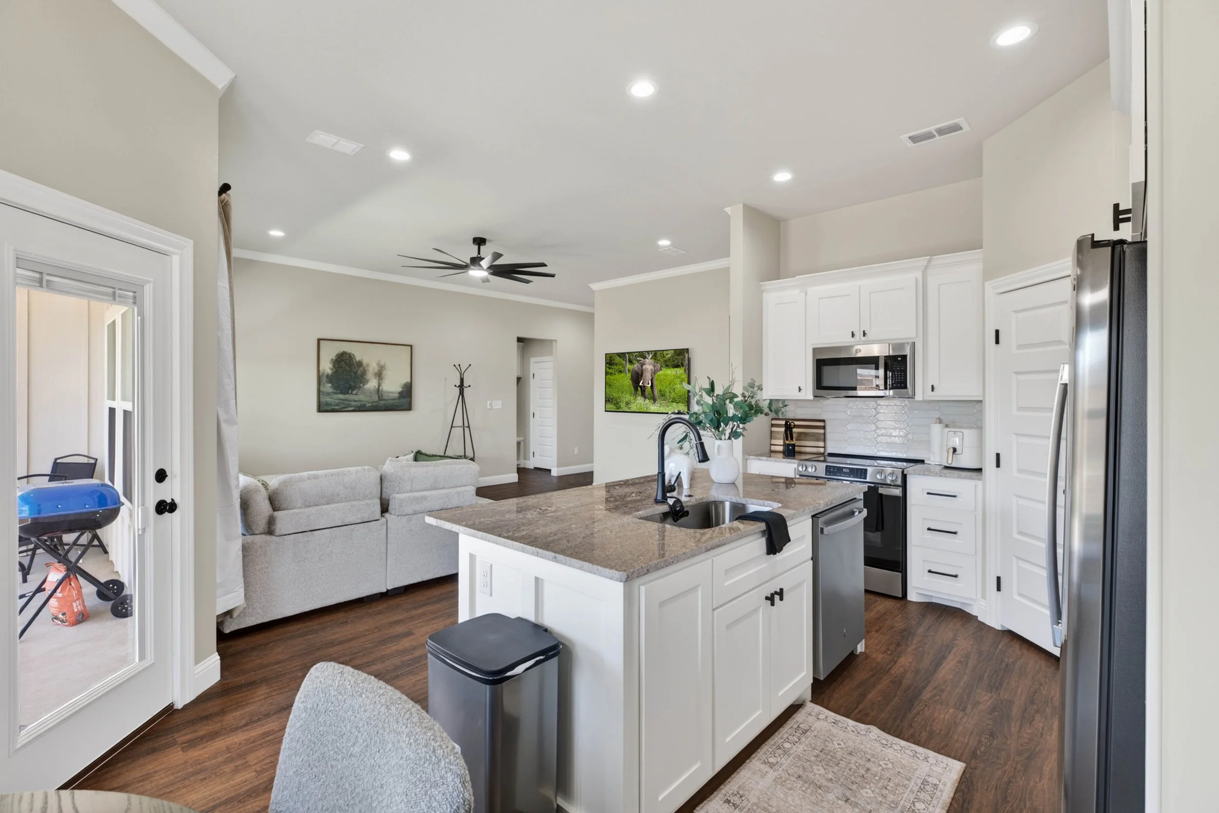 Open-concept kitchen and living room with white cabinets, a granite island, pendant lighting, and a ceiling fan, featuring a wall-mounted TV and decorative artwork