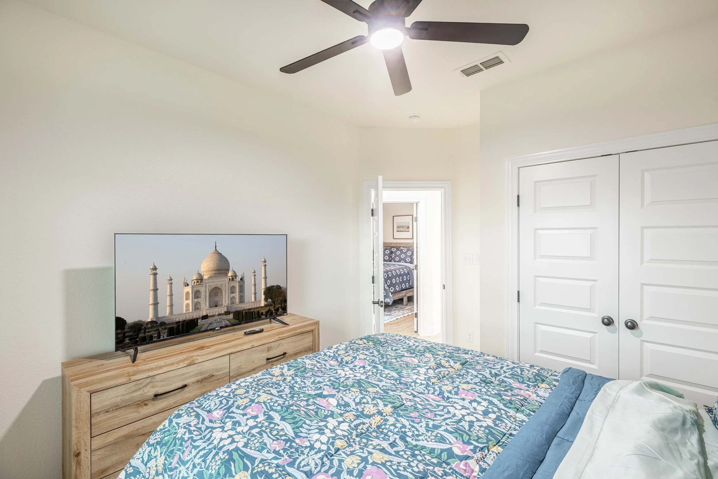 A bedroom with a television displaying the Taj Mahal, a ceiling fan with lights, a doorway leading to another room with a bed, and a closet with white double doors.