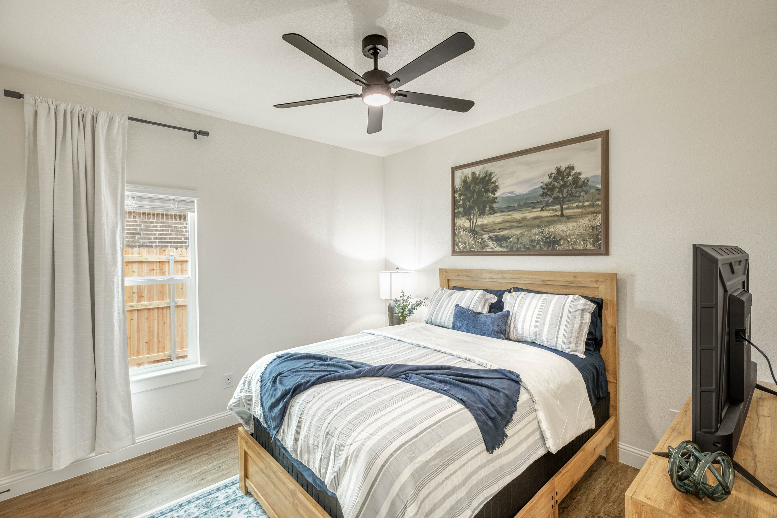 A cozy bedroom with a wooden bed frame, striped bedding, and a landscape painting above the headboard. There is a window with white curtains, a small bedside lamp, and a flat-screen TV on a wooden dresser. A ceiling fan is on the ceiling.