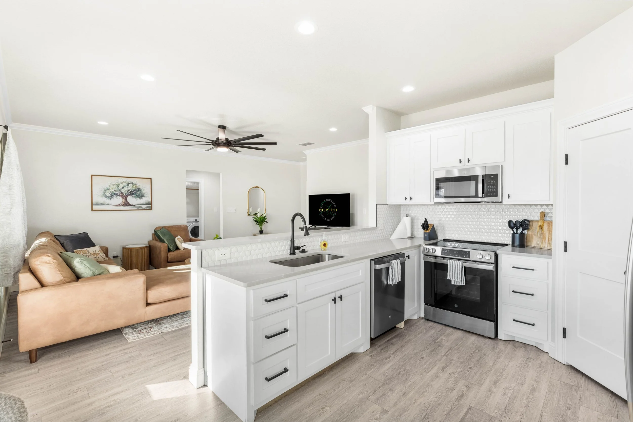 Open-plan living room and kitchen with white cabinetry, stainless steel appliances, and a kitchen island. The living area has a beige sofa with pillows, a wall art of a tree, and a ceiling fan.