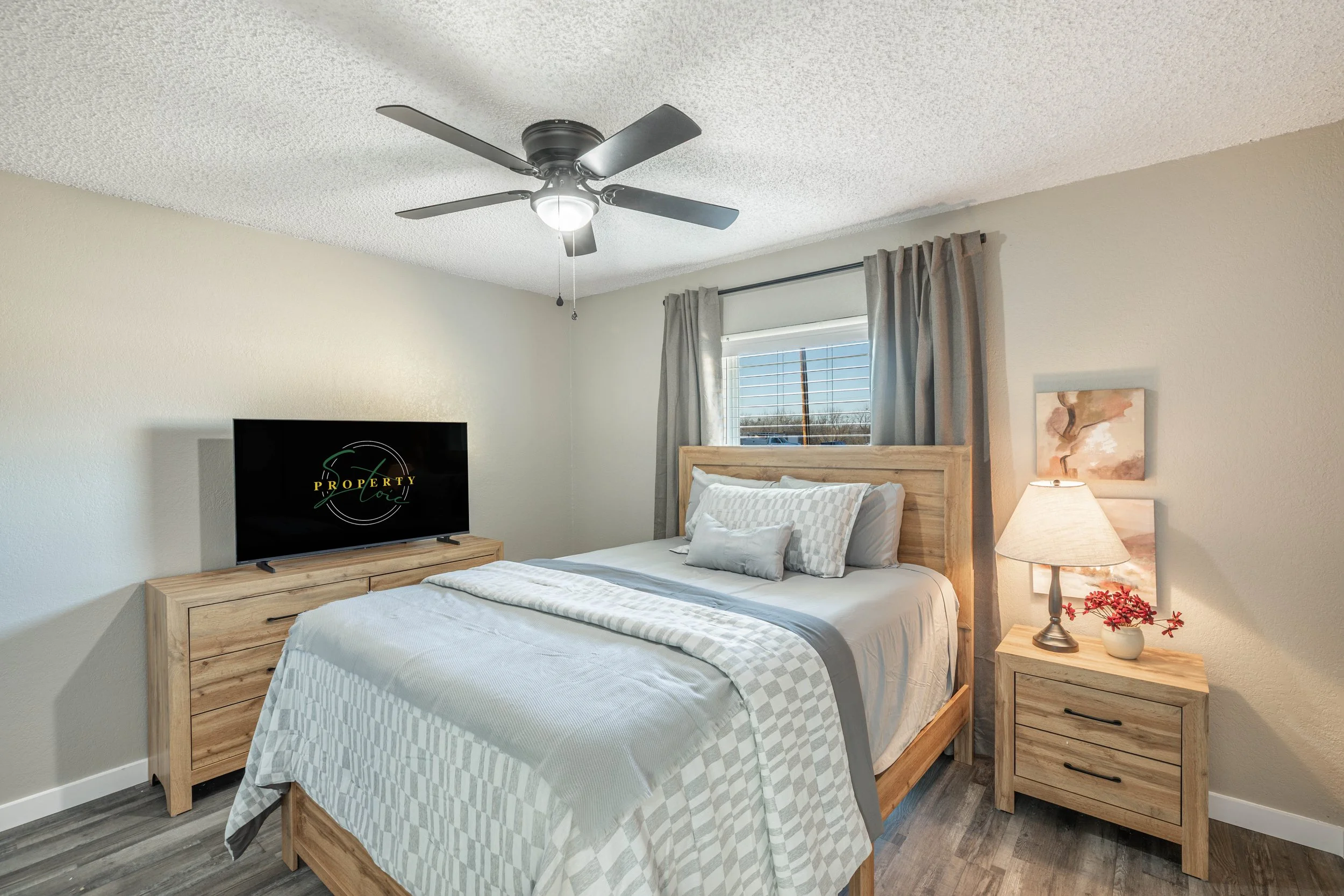 A bedroom with a ceiling fan, a flat-screen TV on a wooden dresser, a bed with a checkered bedspread, a nightstand with a lamp and a small flower arrangement, a window with curtains, and a framed painting on the wall.