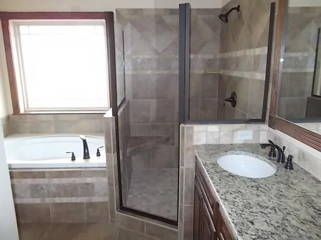 Bathroom with a bathtub next to a window, a walk-in shower with glass doors, and a granite countertop with an oval sink.
