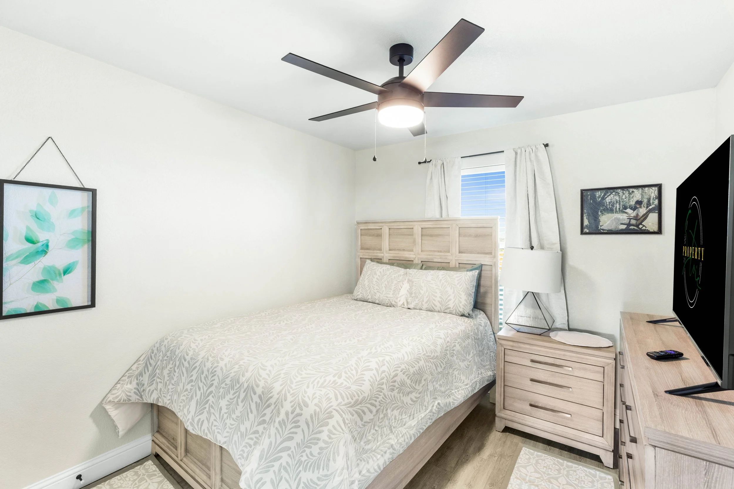 A bedroom with a bed, nightstand, and ceiling fan in a sunny room with white walls and curtains.