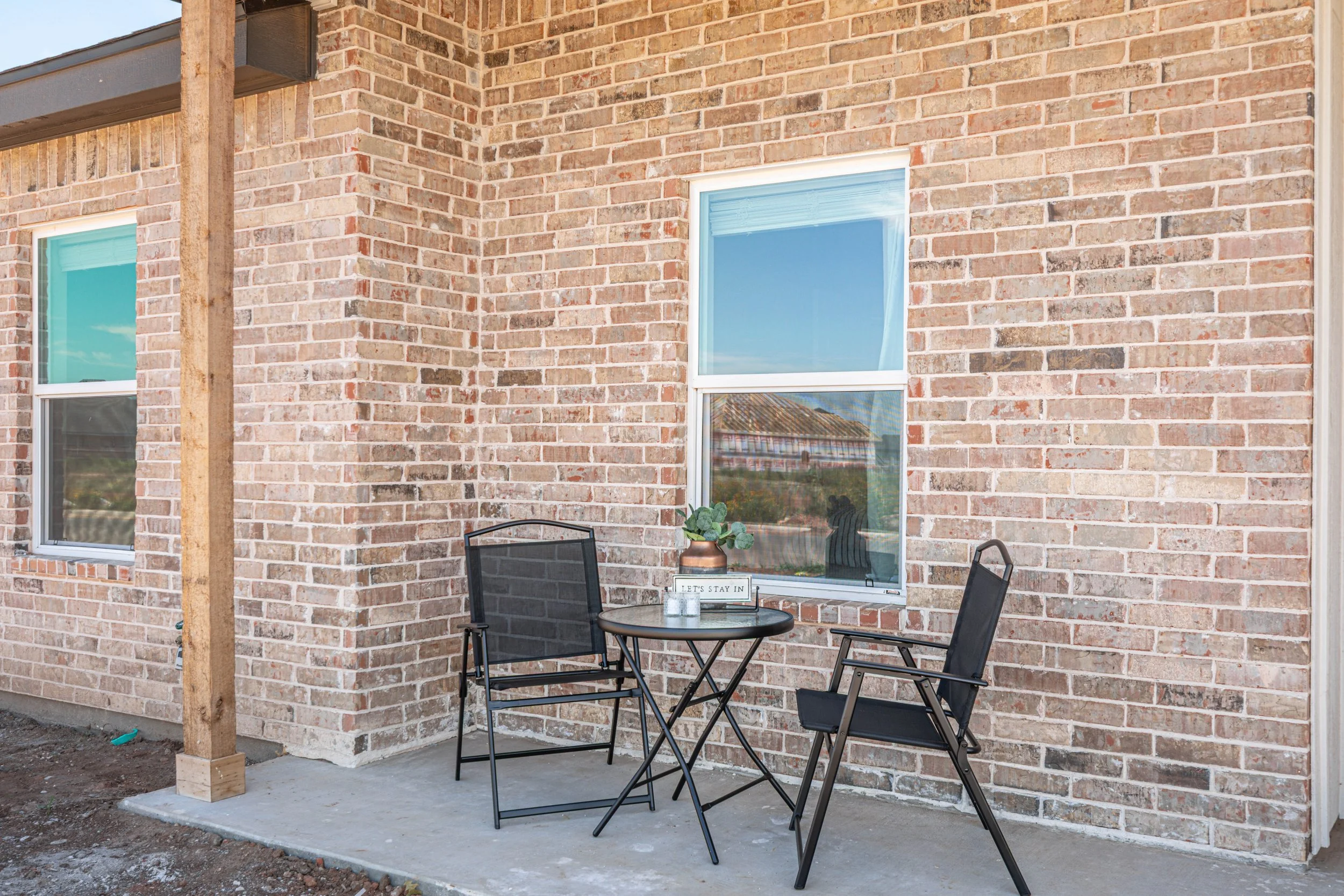 A brick house exterior with two windows and a small patio area featuring a round table and four black chairs. One table has a decorative plant and a sign saying 'Let's Stay In.'