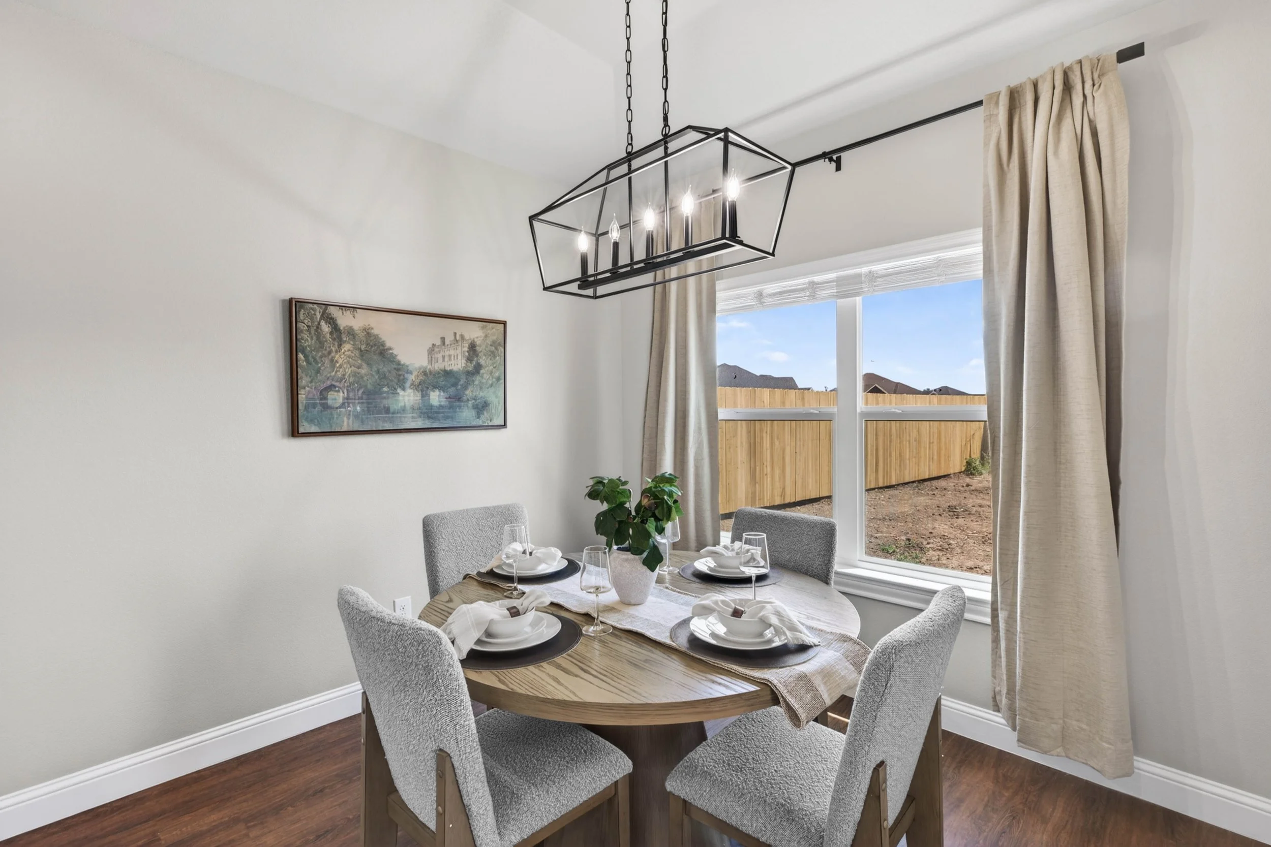 A dining room with a round wooden table set with four place settings, a centerpiece plant, and a beige table runner. Four gray upholstered dining chairs surround the table. A large window with beige curtains and a view of a backyard with a wooden fen