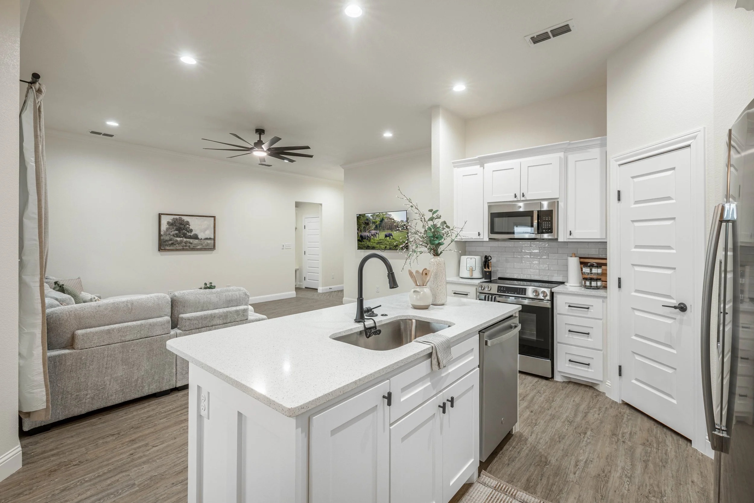 Modern kitchen with white cabinets, stainless steel appliances, and a kitchen island with a sink. Open living space with a ceiling fan and wall art.