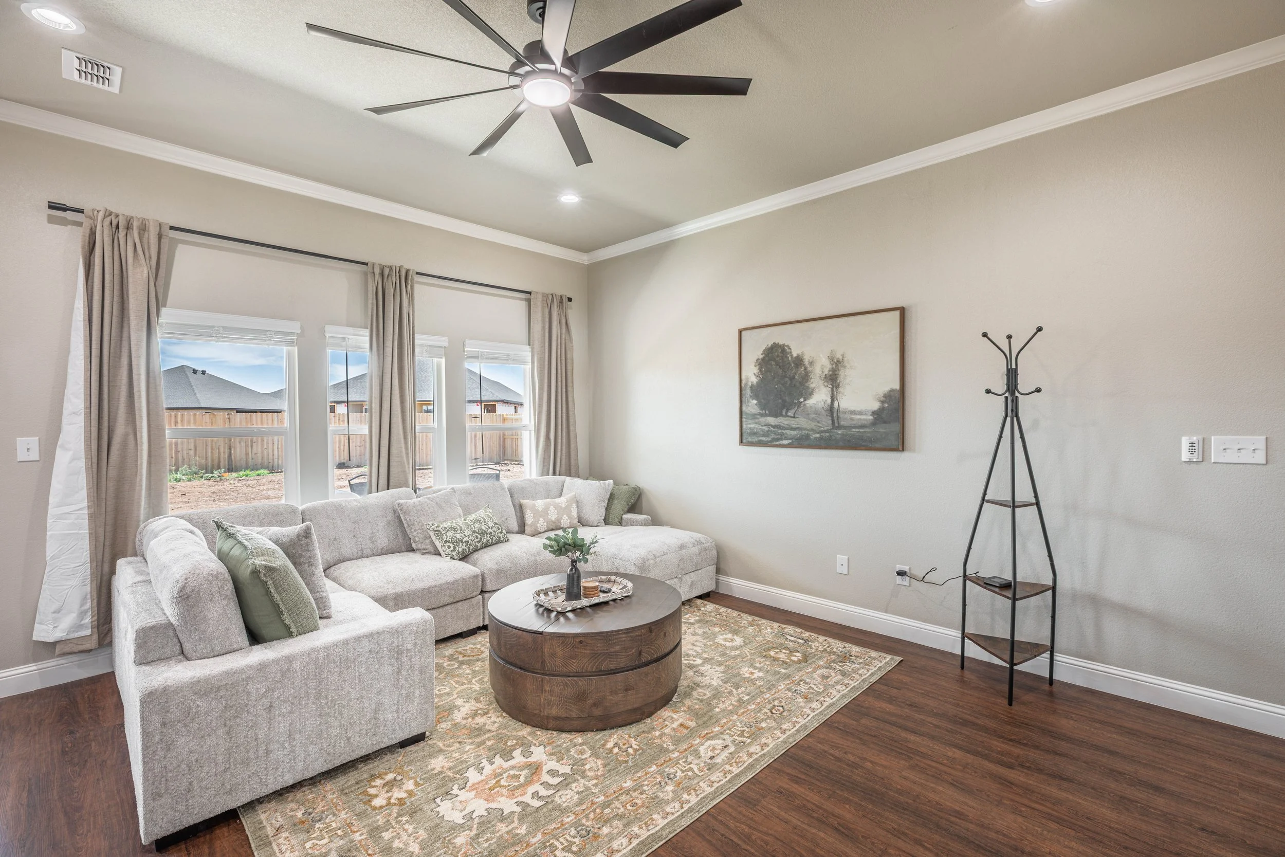 Living room with beige sectional sofa, green pillows, wooden coffee table with tray, large windows with curtains, framed landscape painting, black coat rack, ceiling fan, hardwood floor, patterned area rug.