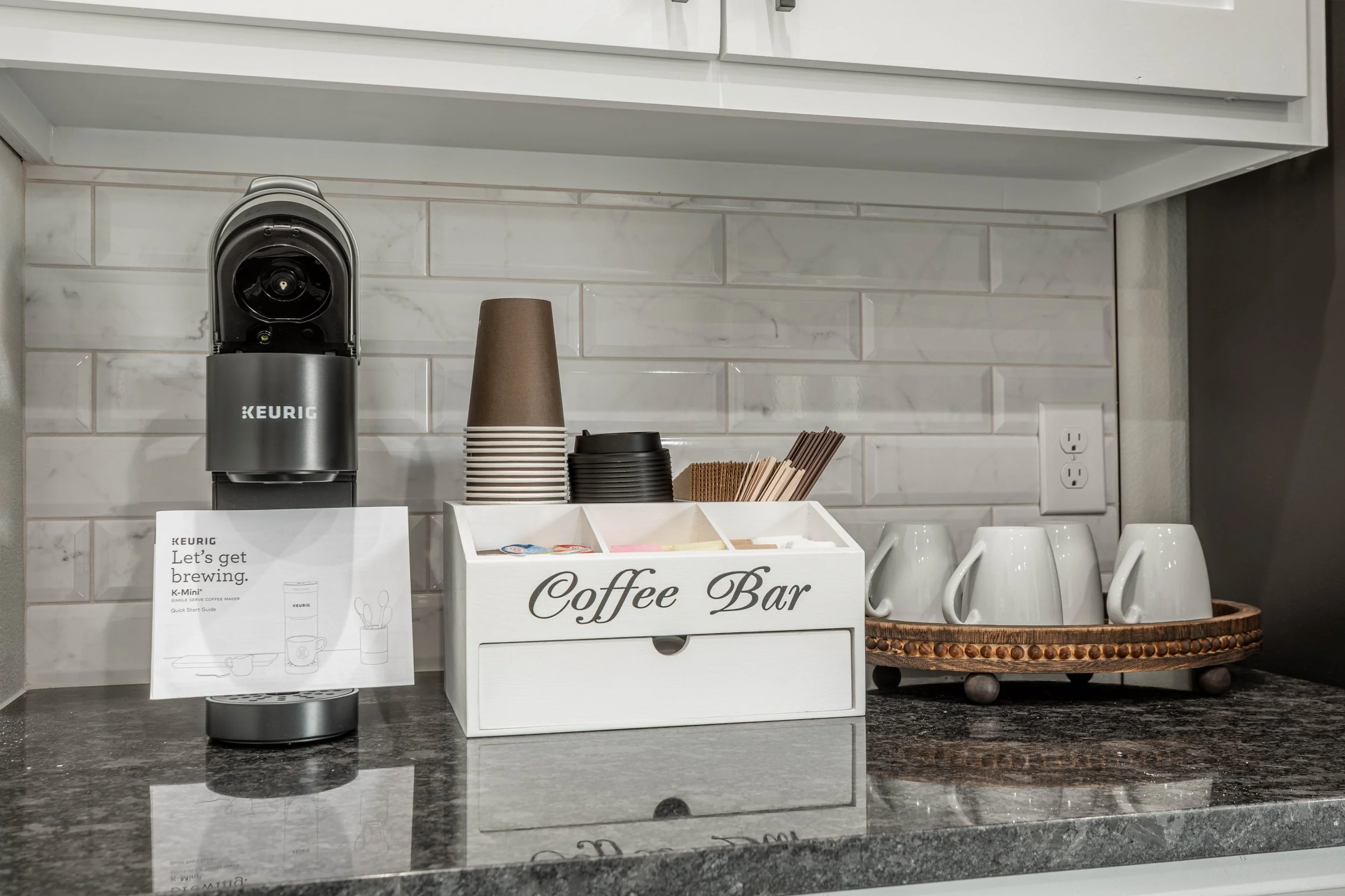 Countertop with a Keurig coffee machine, paper cups, a wooden tray with stir sticks, and a sign that says 'Coffee Bar' in front of a white tiled wall.