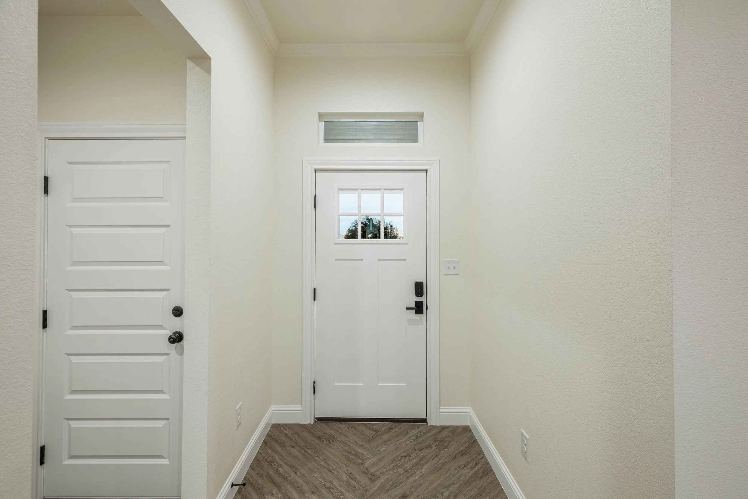 Interior view of the entryway with a white front door featuring four glass panes in the upper half and a black handle. To the left is a white door with horizontal paneling and a black doorknob. The walls are painted light cream, and the flooring is w
