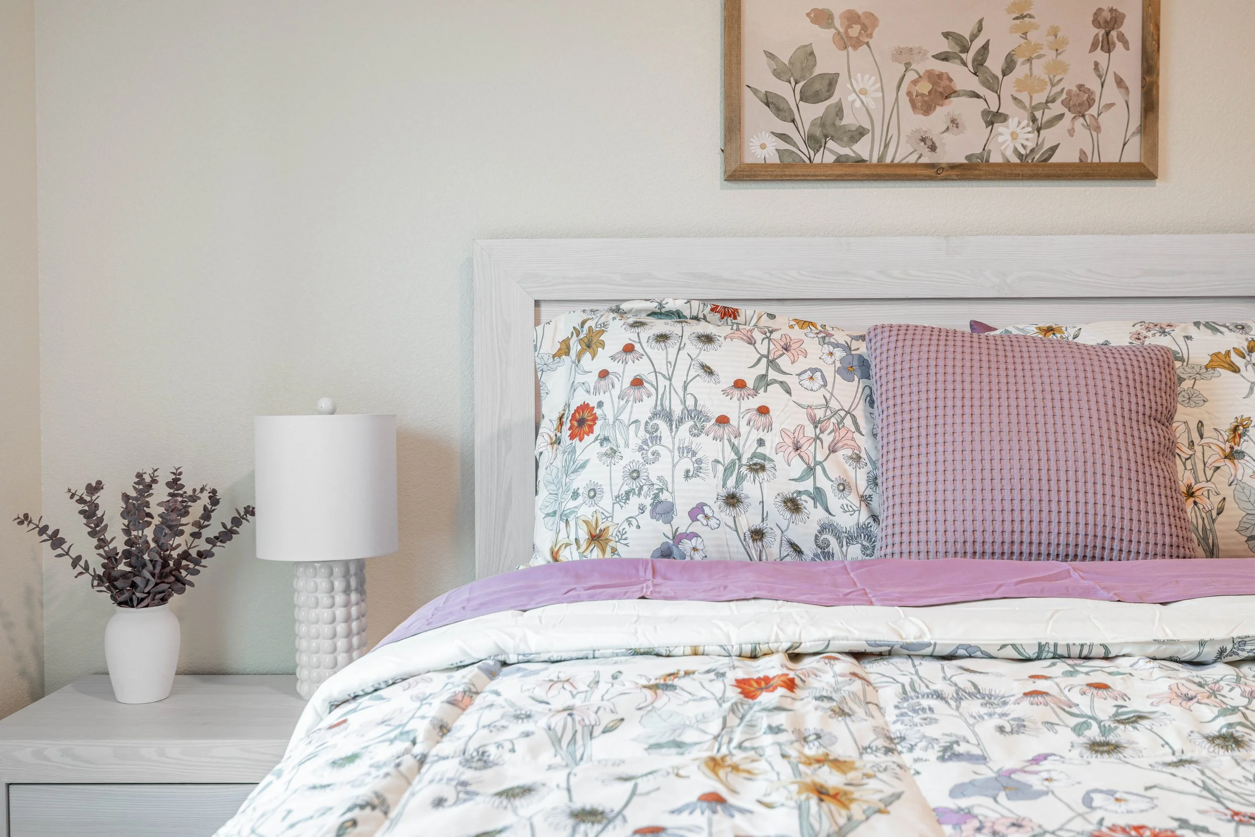 A bedroom with a white bed frame, floral bedding, and a pink pillow. Next to the bed, there's a white nightstand with a round white lamp and a white vase with dark purple branches. A framed floral painting hangs on the off-white wall above.