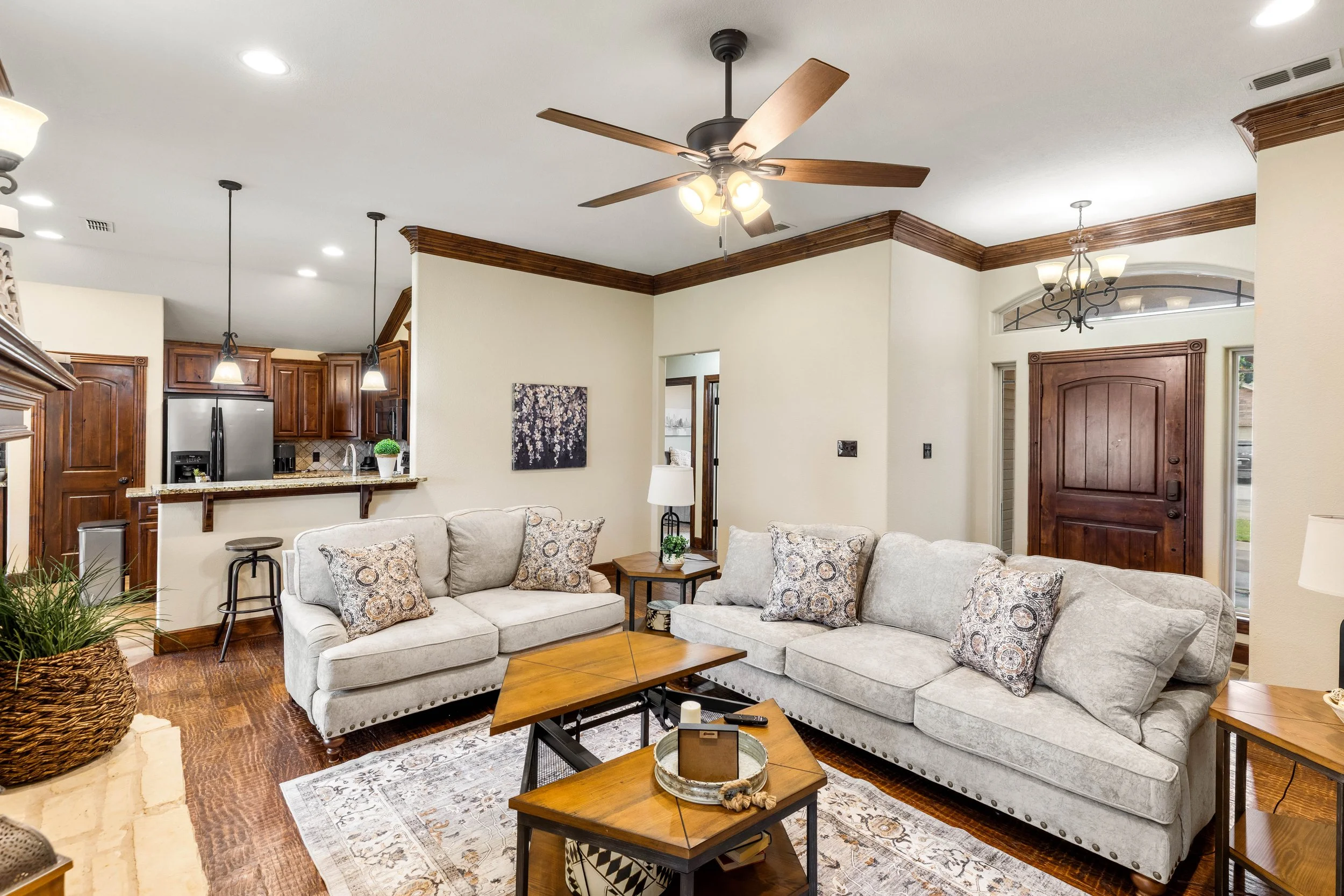 Living room with beige sofas, patterned cushions, wooden coffee tables, and a rug. Open kitchen area with wooden cabinets, stainless steel refrigerator, and pendant lights. Ceiling fan with lights. Entry door with arch and windows above.