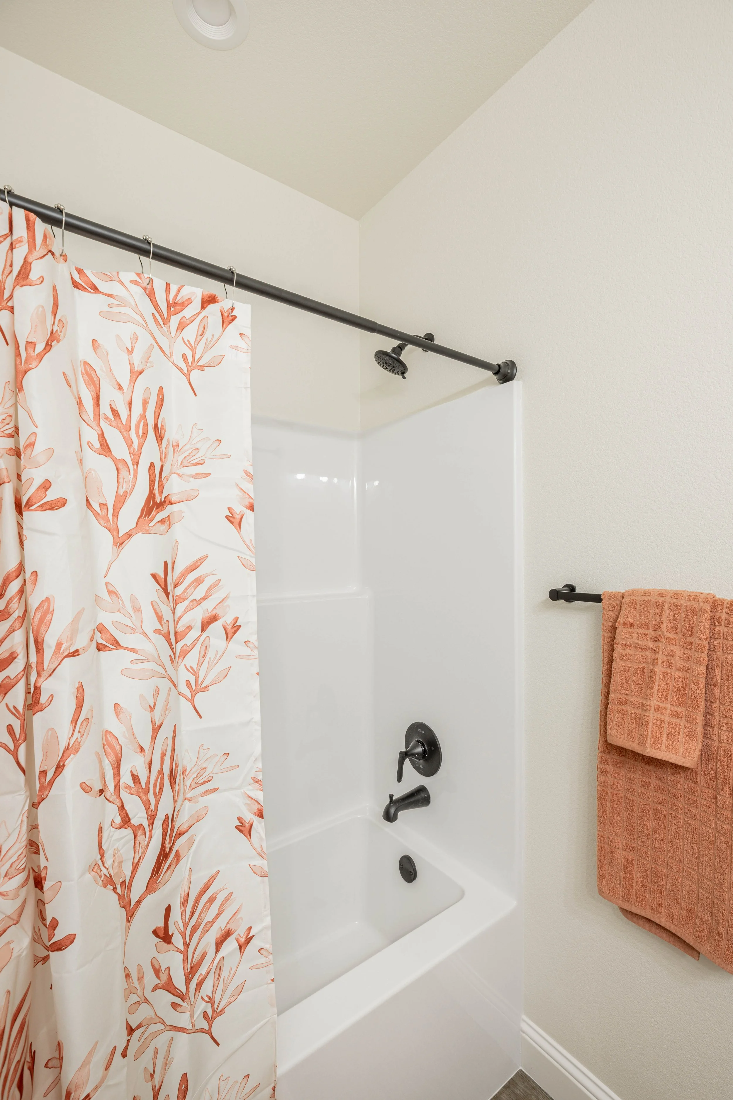 Bathroom shower with a coral-patterned shower curtain, white tub, black fixtures, orange towels on a black towel bar, and a round ceiling light.