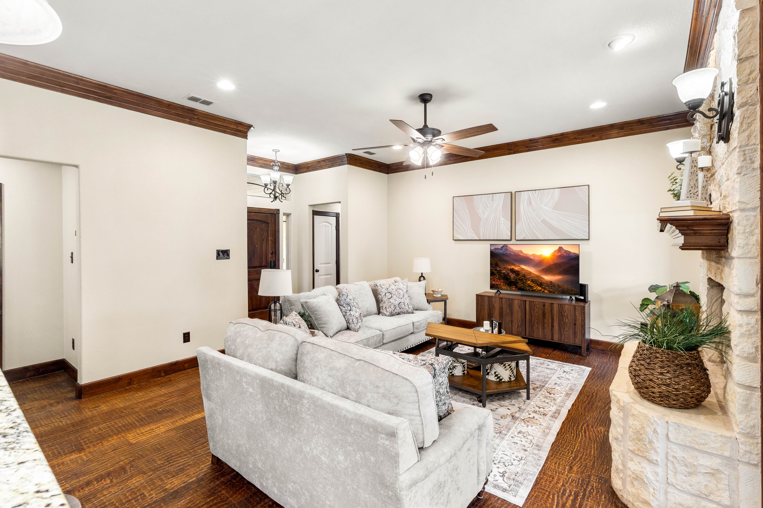 A living room with a white sofa, patterned cushions, a wooden coffee table, a television on a wooden stand, wall art, a ceiling fan, and wall-mounted lights.