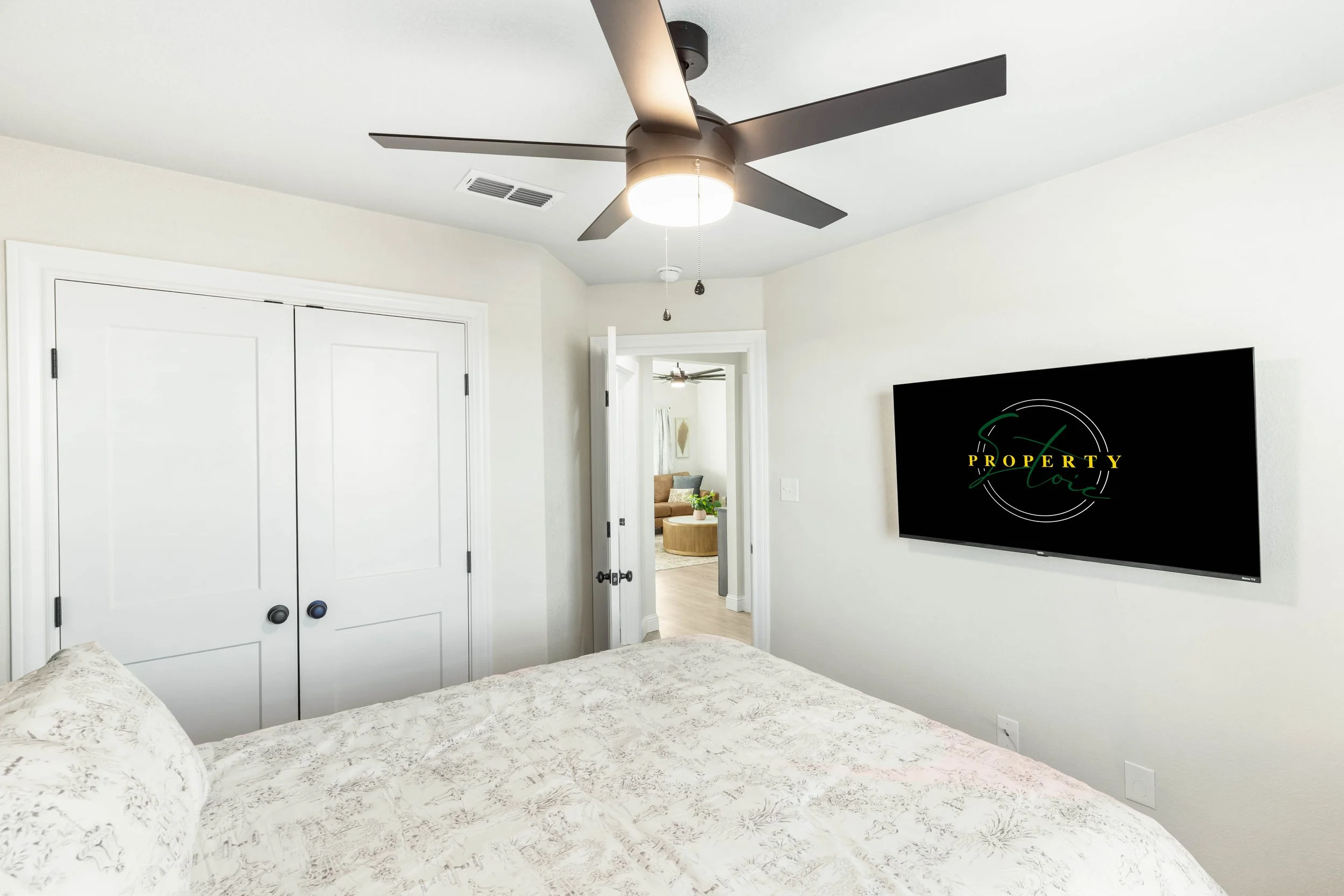 A bedroom with a ceiling fan, a wall-mounted TV, a closet with double doors, and a view into another living space with a sofa and armchair.