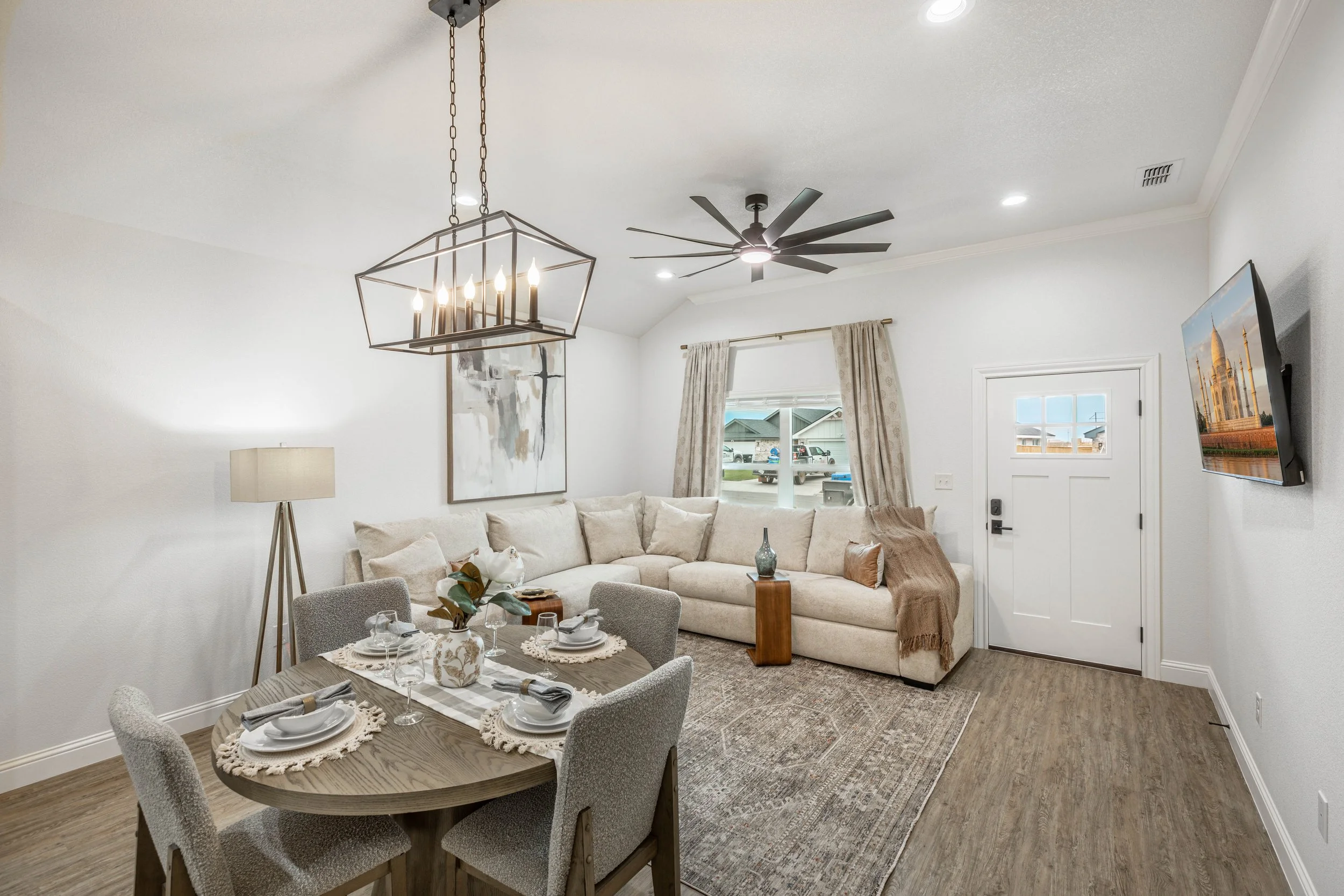 Living room with a beige sectional sofa, a round dining table set with tableware, a modern chandelier, a ceiling fan, a window with beige curtains, a wall-mounted TV, and artwork on the wall.