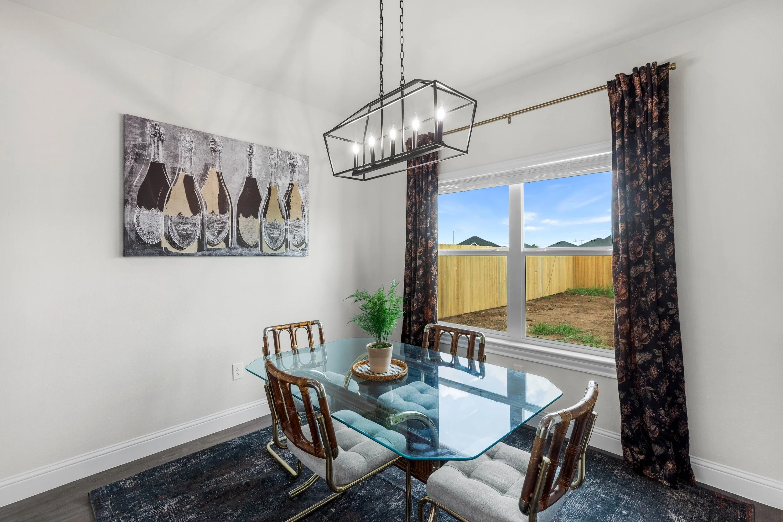 A dining room with a glass table, four chairs with cushioned seats, a red potted plant, a modern rectangular chandelier, a large artwork of beer bottles on the wall, and a window with floral curtains showing a backyard with a wooden fence and blue sk