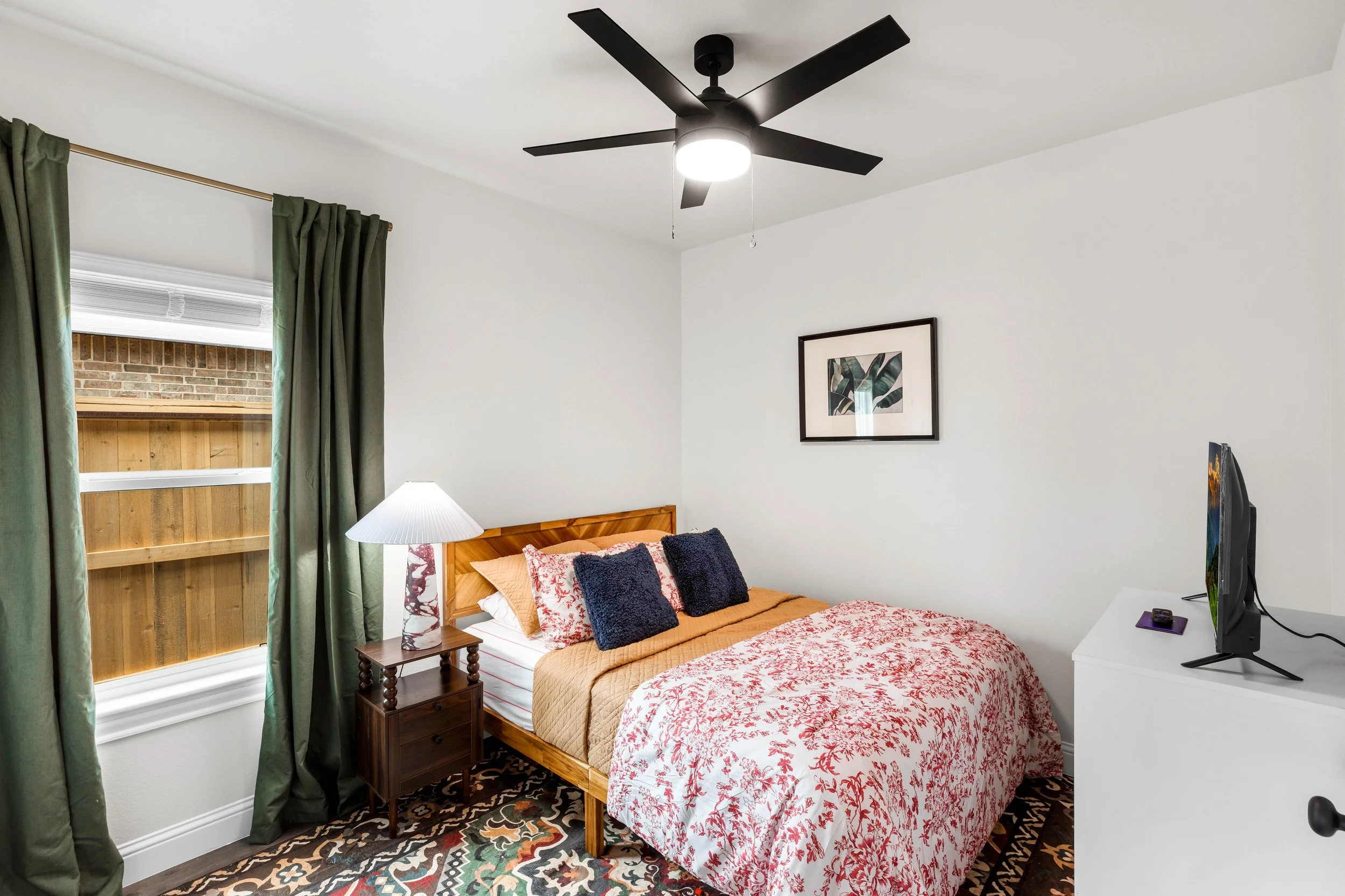 A bedroom with a bed covered in a red and white floral quilt, three pillows, a wooden nightstand with a lamp, a window with green curtains, a framed art piece on the wall, a TV on a white stand, and a ceiling fan with lights.