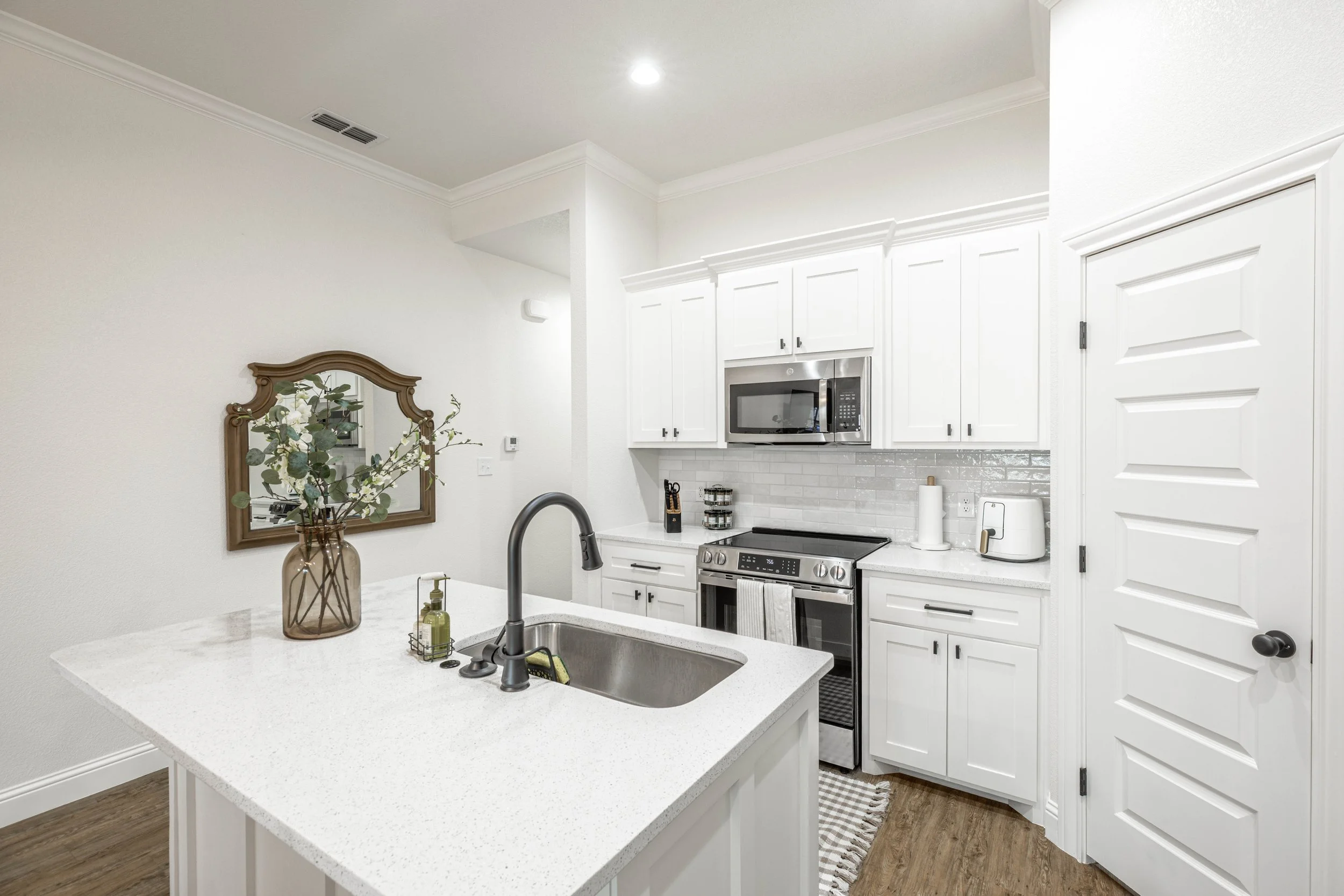 Modern white kitchen with granite countertop island, stainless steel appliances, and white cabinets. Decor includes a vase with flowers, soap dispensers, and a paper towel holder. There is a mirror and a door visible in the background.
