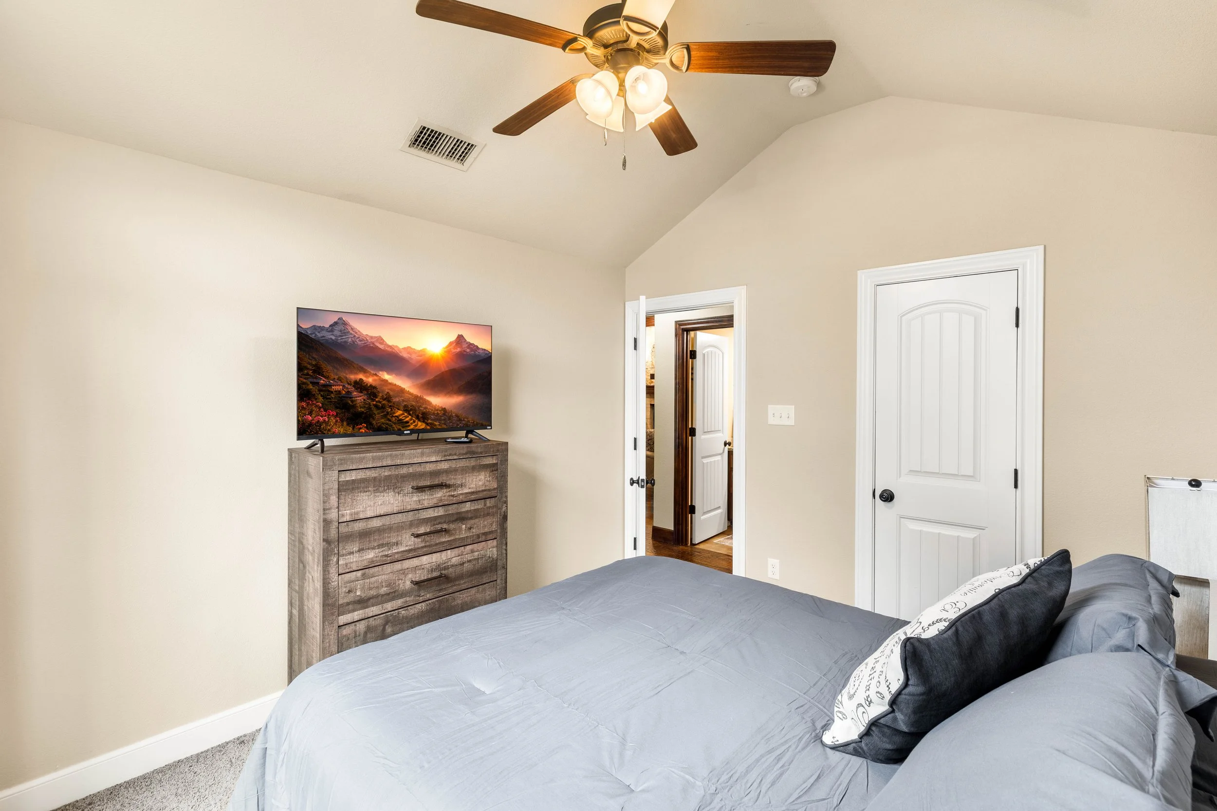 A bedroom with beige walls, a ceiling fan with attached lights, a dresser with a TV displaying a mountain landscape, and three doors, one slightly open showing a glimpse of another room.