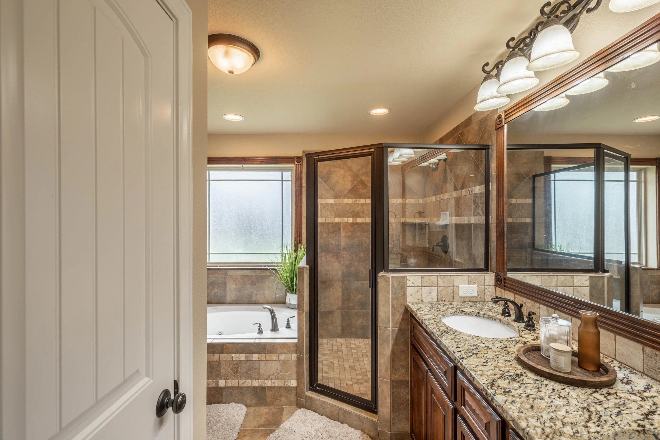Bathroom with a granite countertop vanity, a framed mirror, a glass-enclosed shower, a bathtub, a window, and ceiling lights.