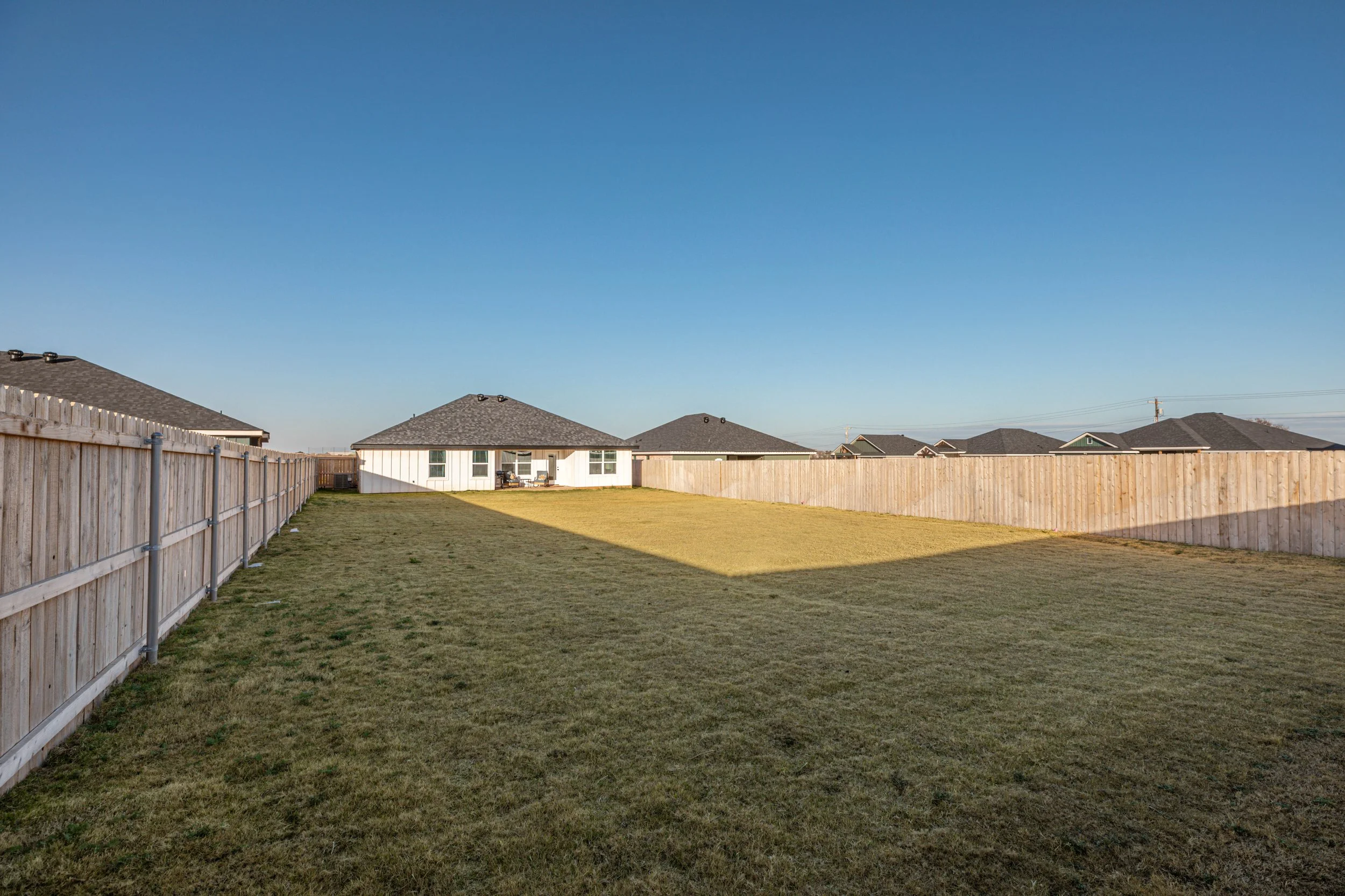 Backyard with a grassy lawn, wooden fence, and houses with gray roofs under a clear blue sky.