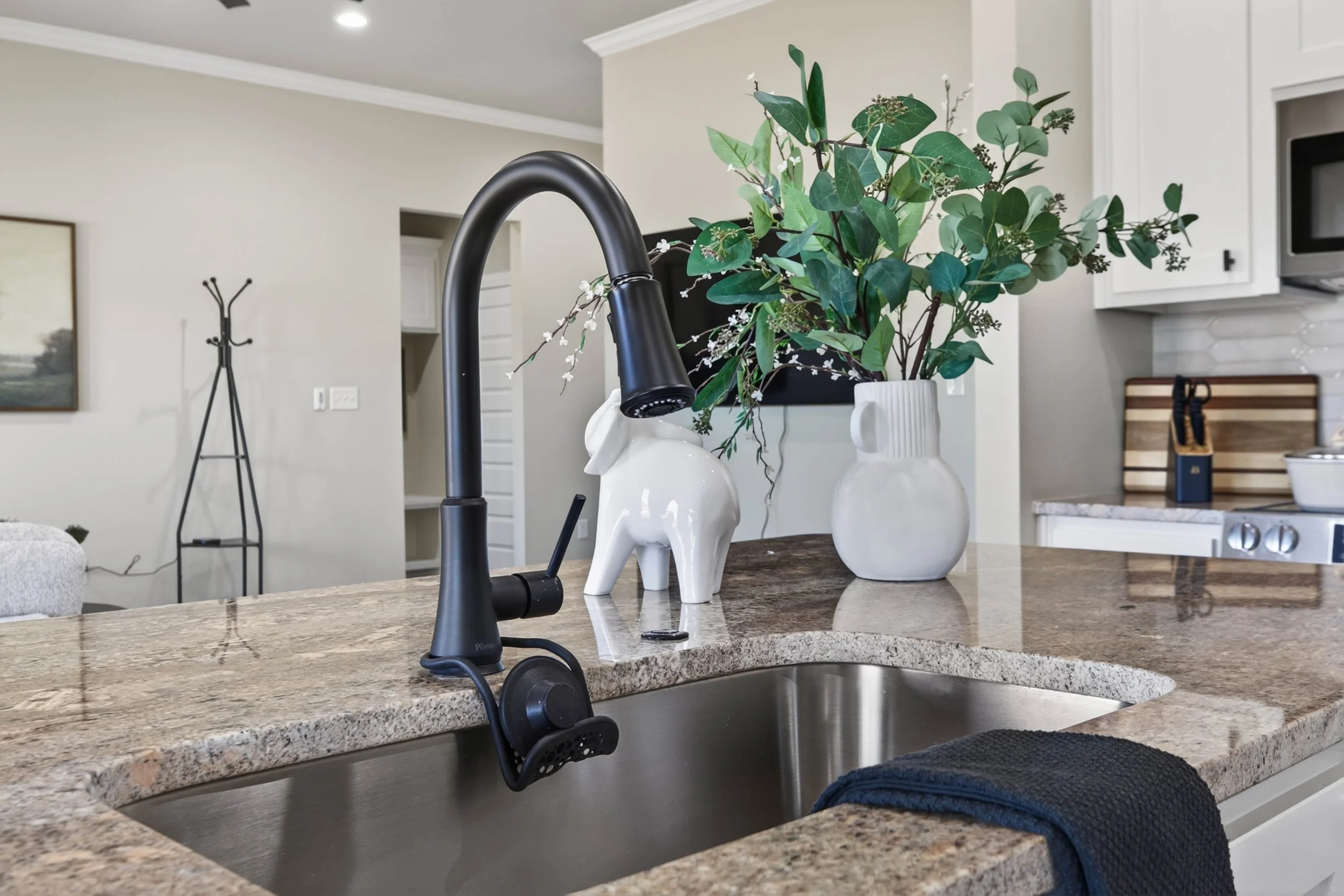 A kitchen countertop with a black faucet, two white vases, and a potted plant. The background includes a living area with a coat rack, artwork on the wall, and kitchen cabinets.
