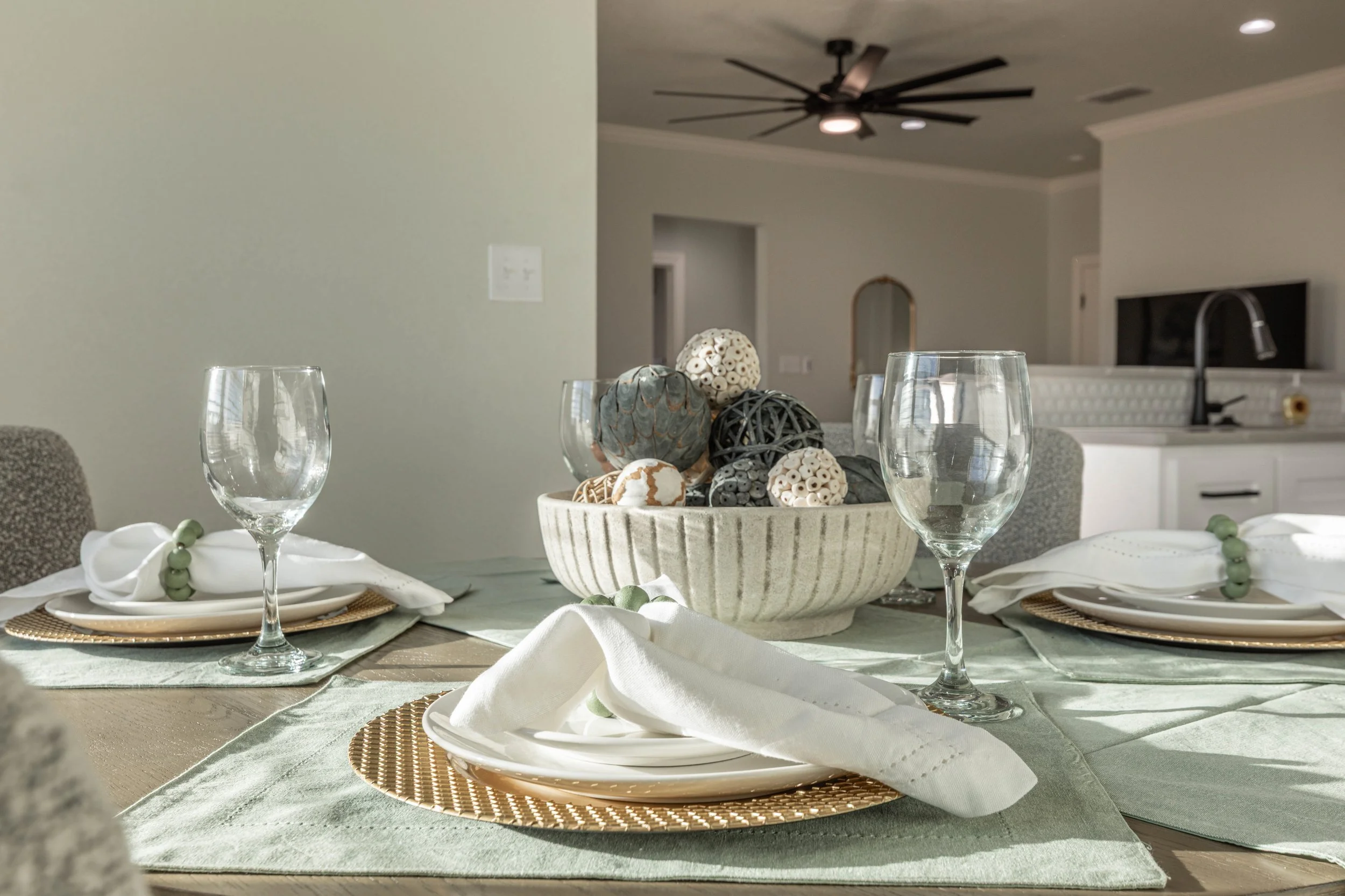 A dining table set with white plates, gold charger plates, white napkins with green bead napkin rings, and empty wine glasses. The center of the table has a large bowl filled with decorative objects that look like natural pods and balls. The backgrou