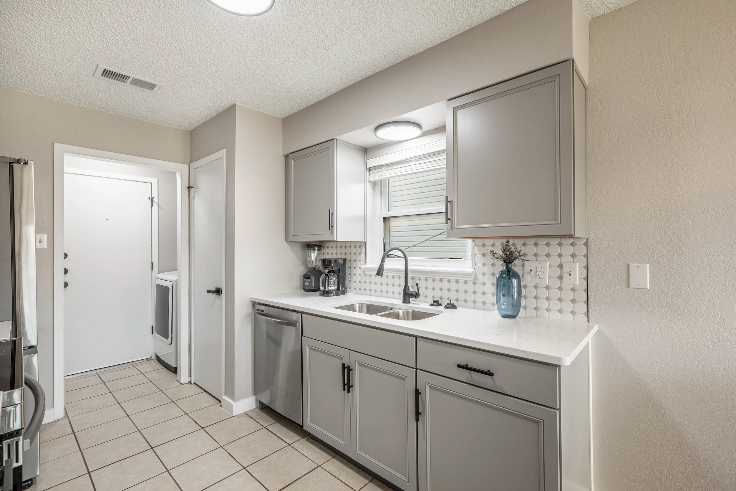 A modern kitchen with gray cabinets, a white countertop, a stainless steel sink, a window with blinds, and a potted plant in a blue vase. There are ceiling lights and tiled flooring.