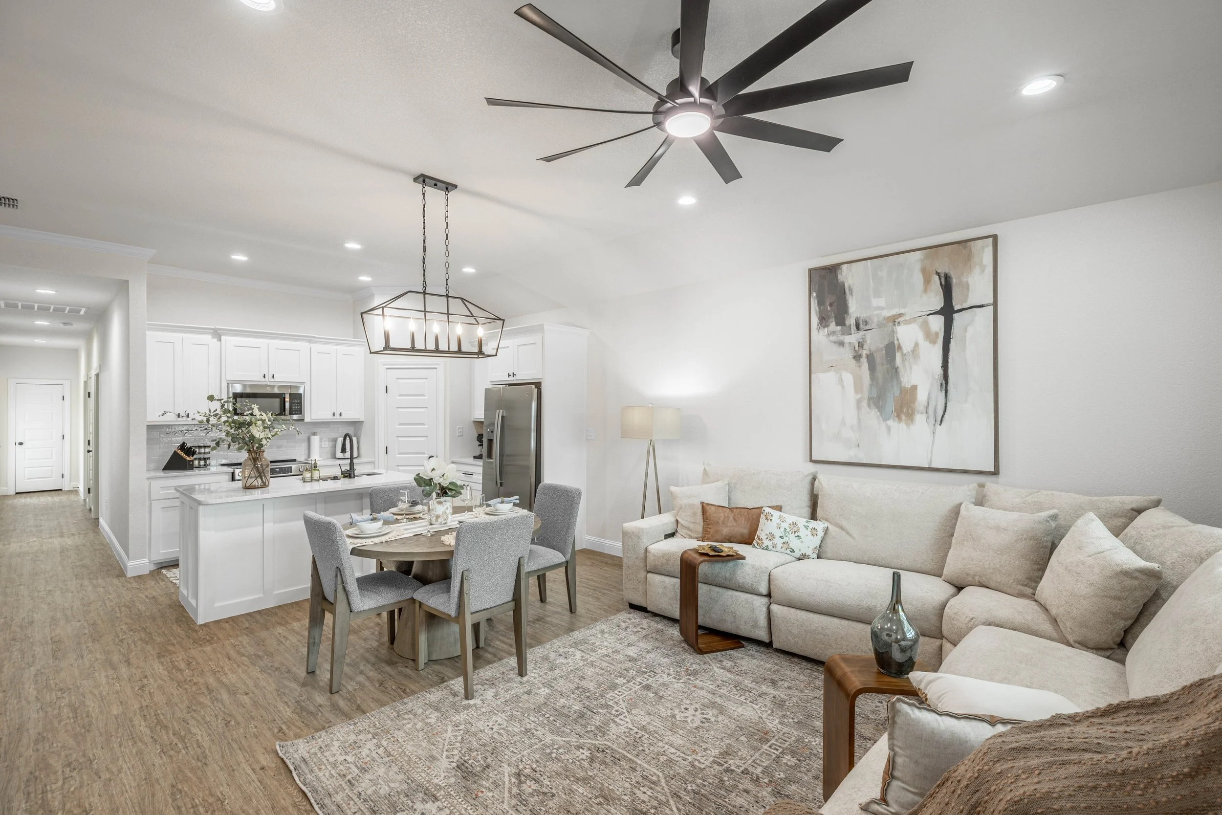 Open-concept living area with white kitchen, dining table, beige sofa, large abstract painting, ceiling fan, and hardwood floors.
