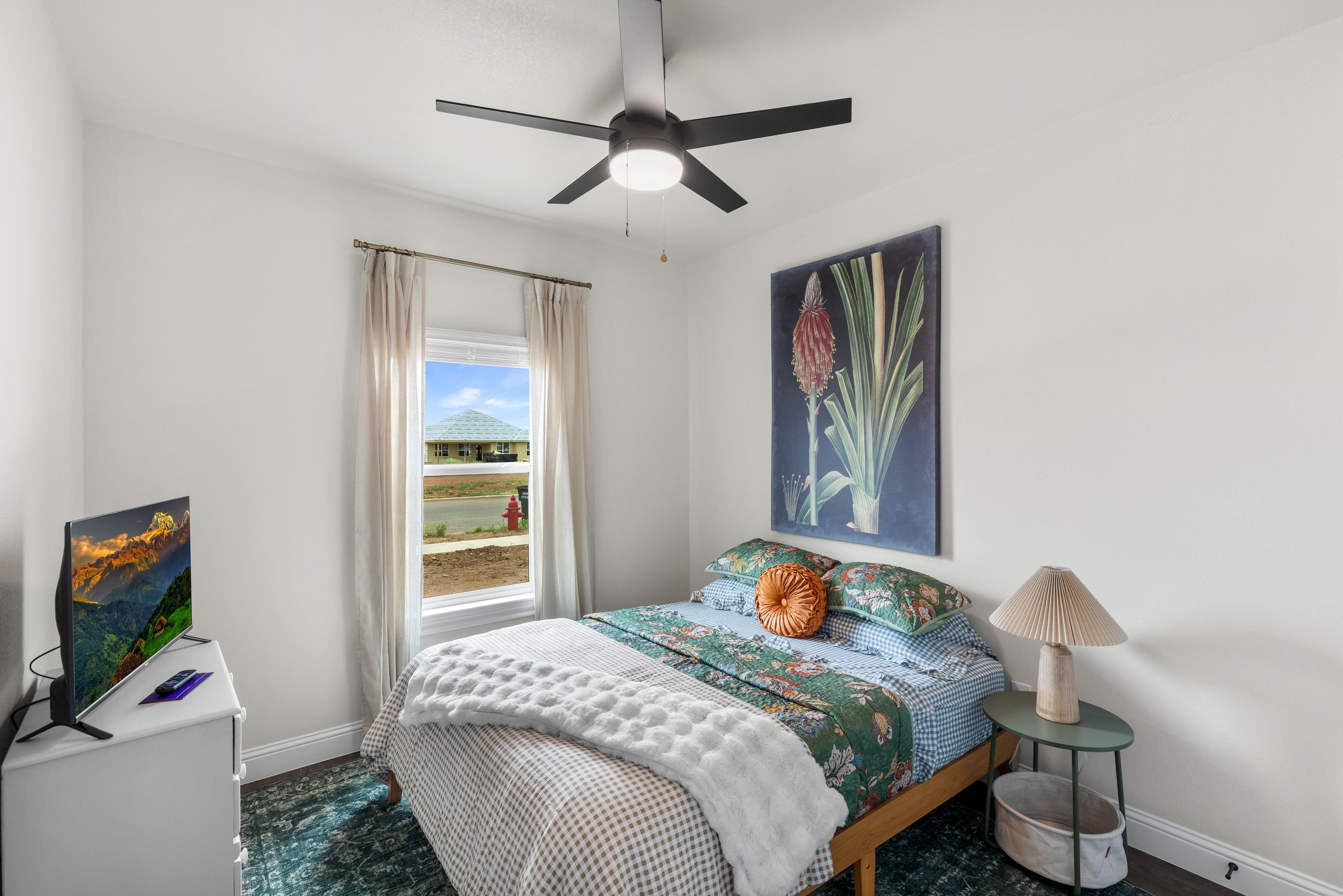 A bedroom with a bed, a small side table with a lamp, a window with cream curtains, a wall hanging of a plant, and a TV showing mountains and sunrise.
