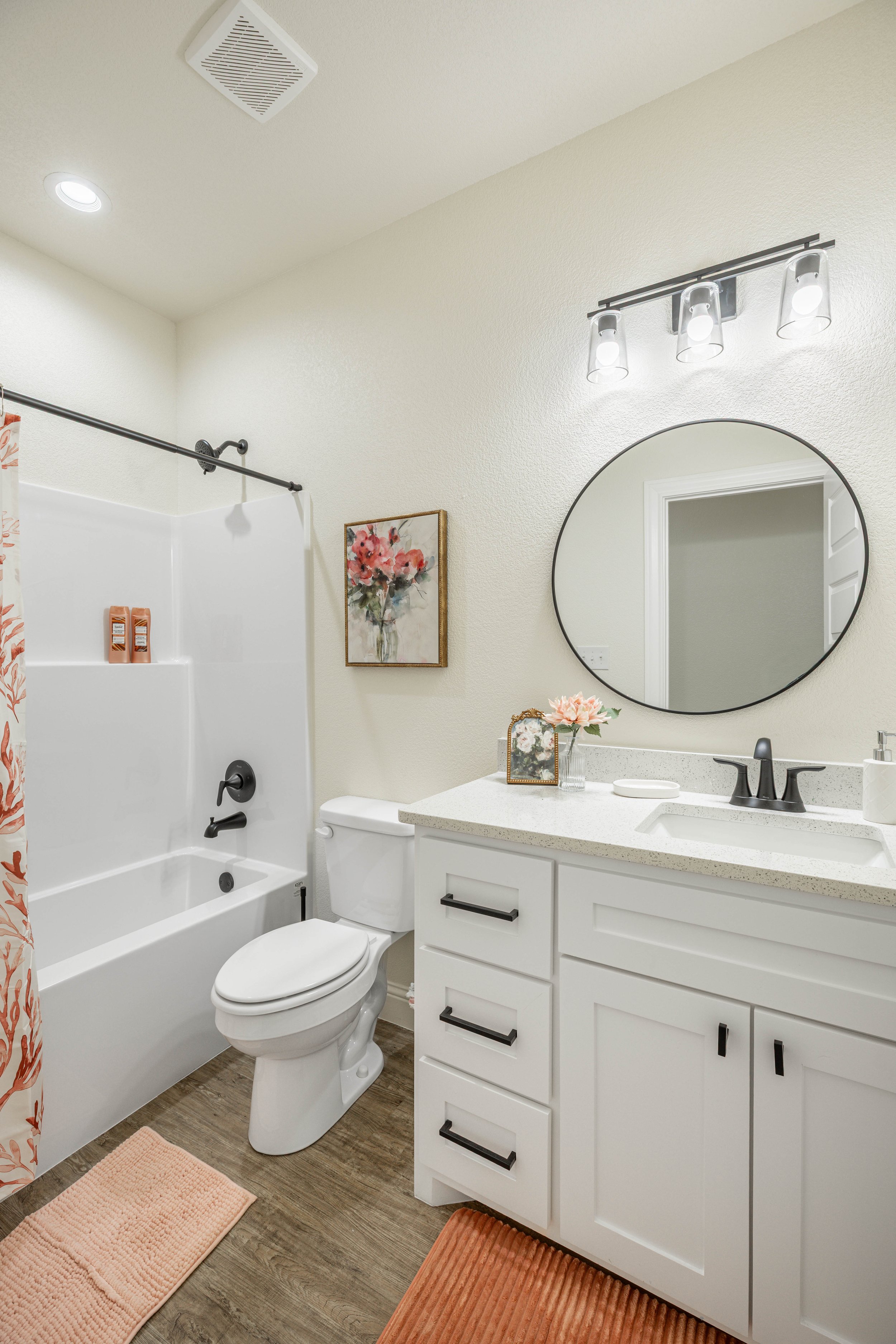A clean and modern bathroom featuring a shower and bathtub with a floral shower curtain, a toilet, a white vanity with black handles, a round mirror, floral decorations, and orange rugs on wood-look flooring.