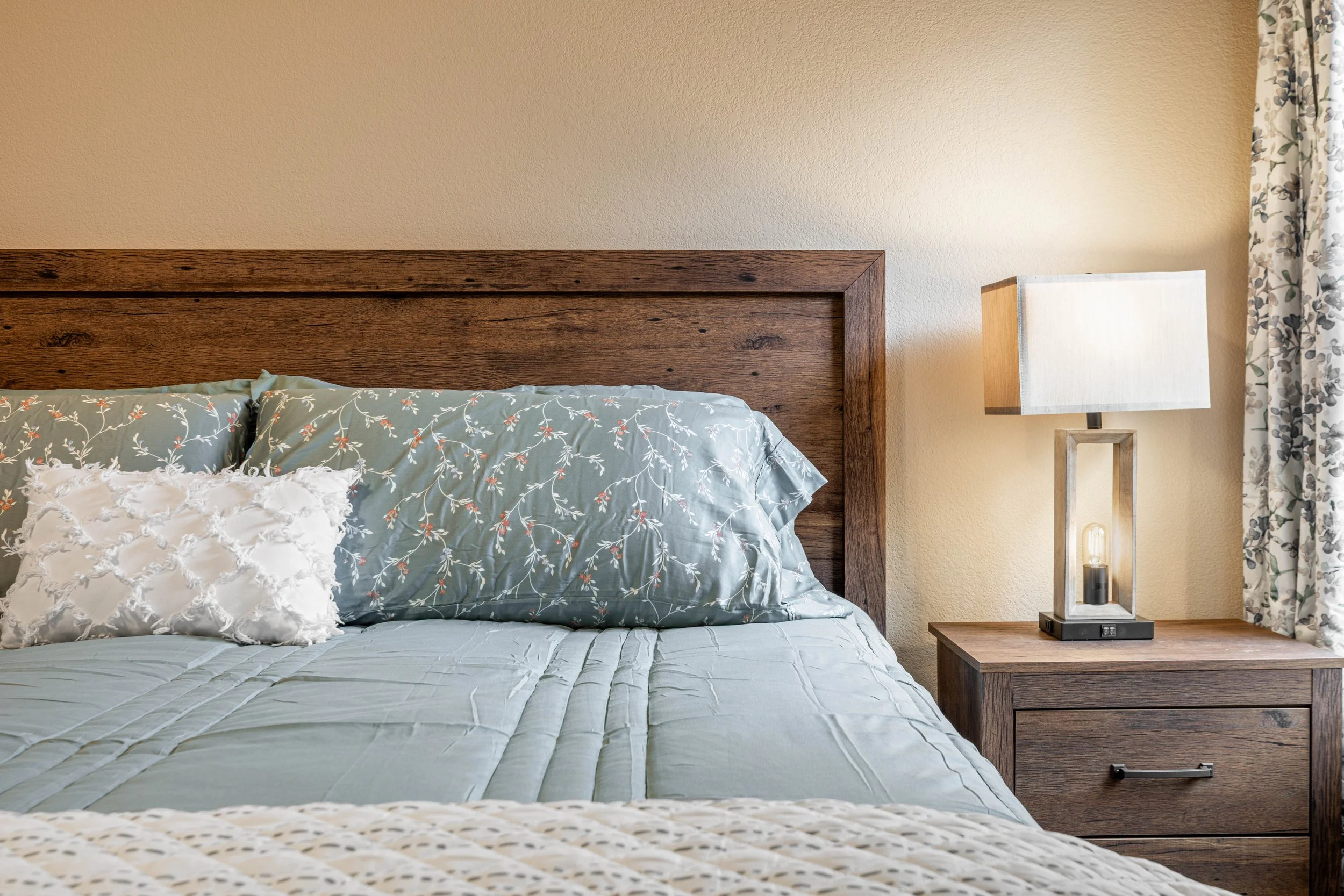 A bedroom with a wooden headboard, floral bedding, and a nightstand with a modern lamp against a beige wall.