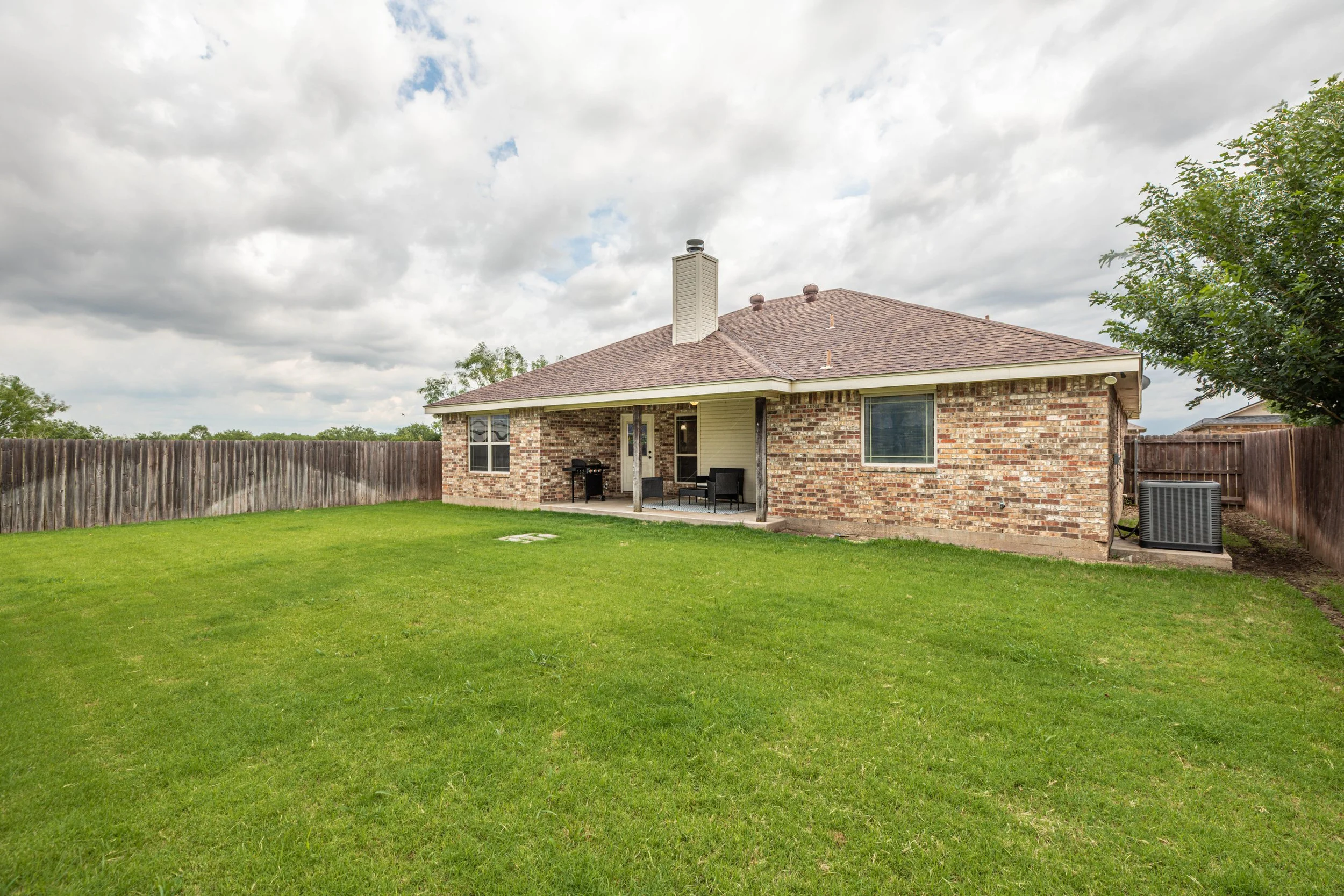 Backyard view of a brick house with a covered patio, green lawn, wooden fence, and cloudy sky.