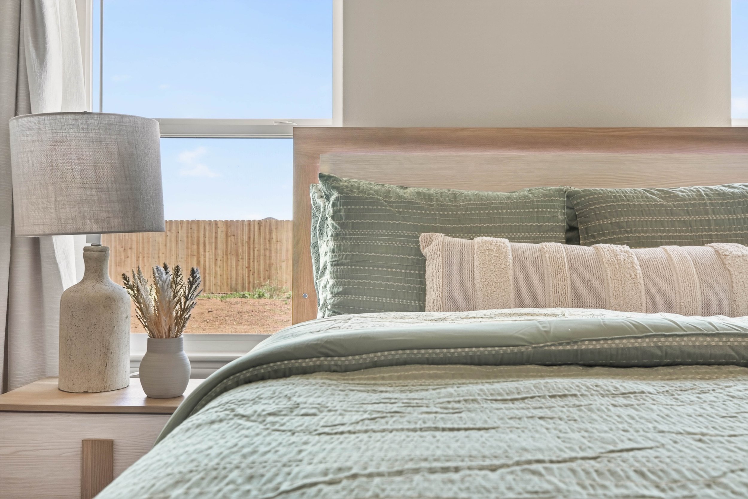 A bedroom with beige bedding, a white bedside table with two decorative vases, a table lamp, a window with a view of a wooden fence and blue sky, and part of a wood headboard.