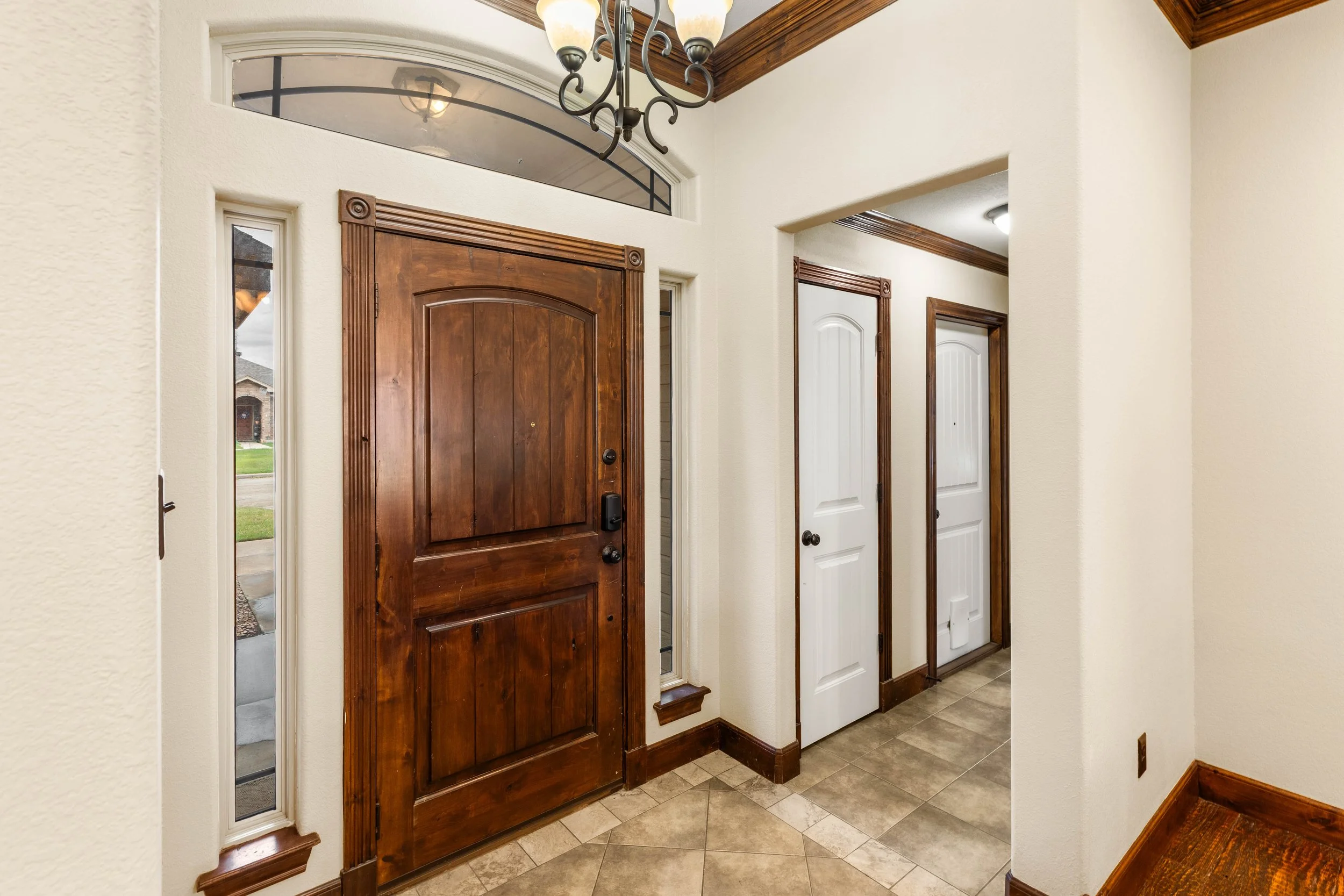The image shows the interior of a house entryway with multiple doors, including a prominent wooden front door with a rounded top and a black door handle. The entryway has beige textured walls, a tiled floor, wooden trim, and a ceiling with a hanging 