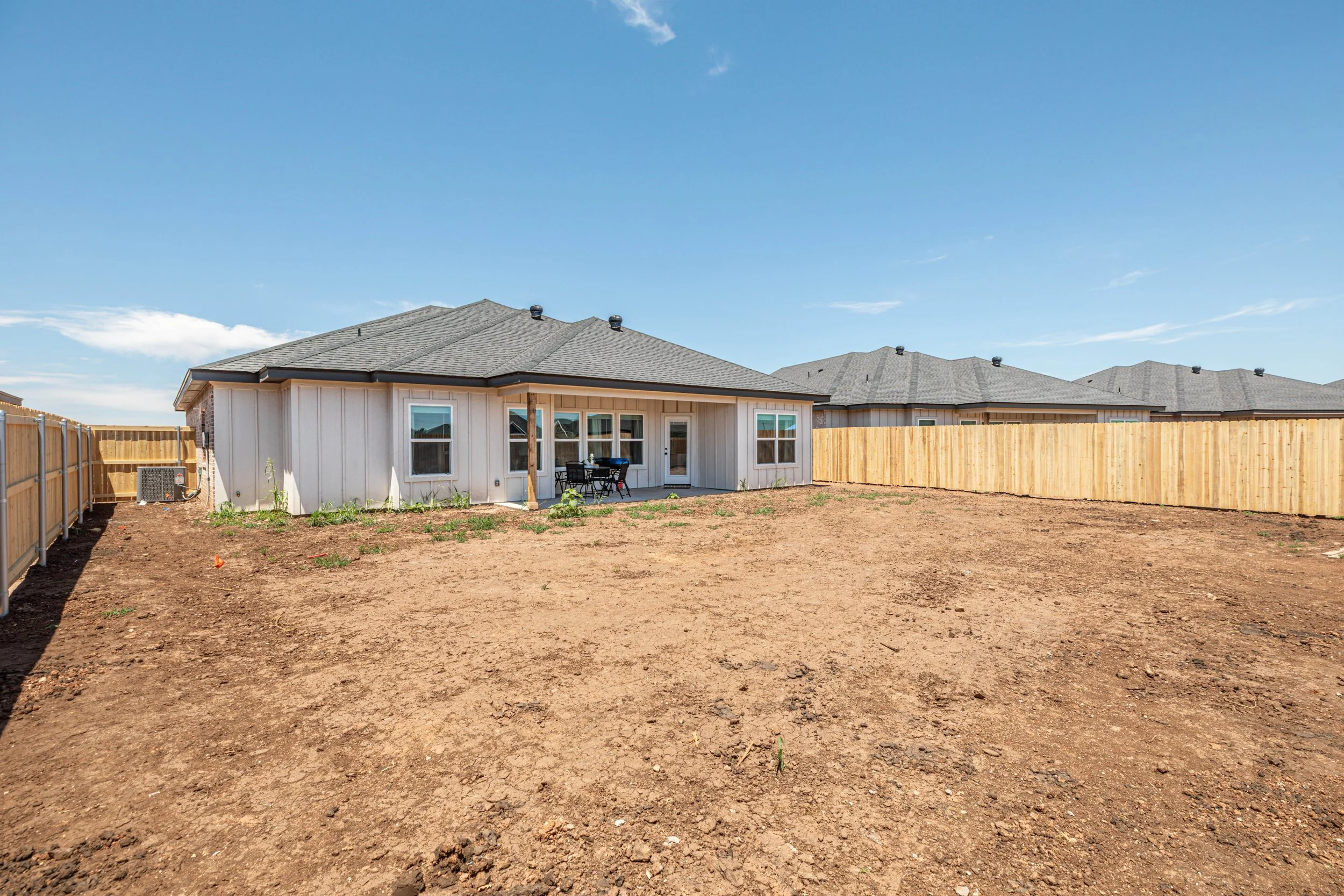Backyard with a newly built house, fenced backyard, and dirt ground, under a clear blue sky.