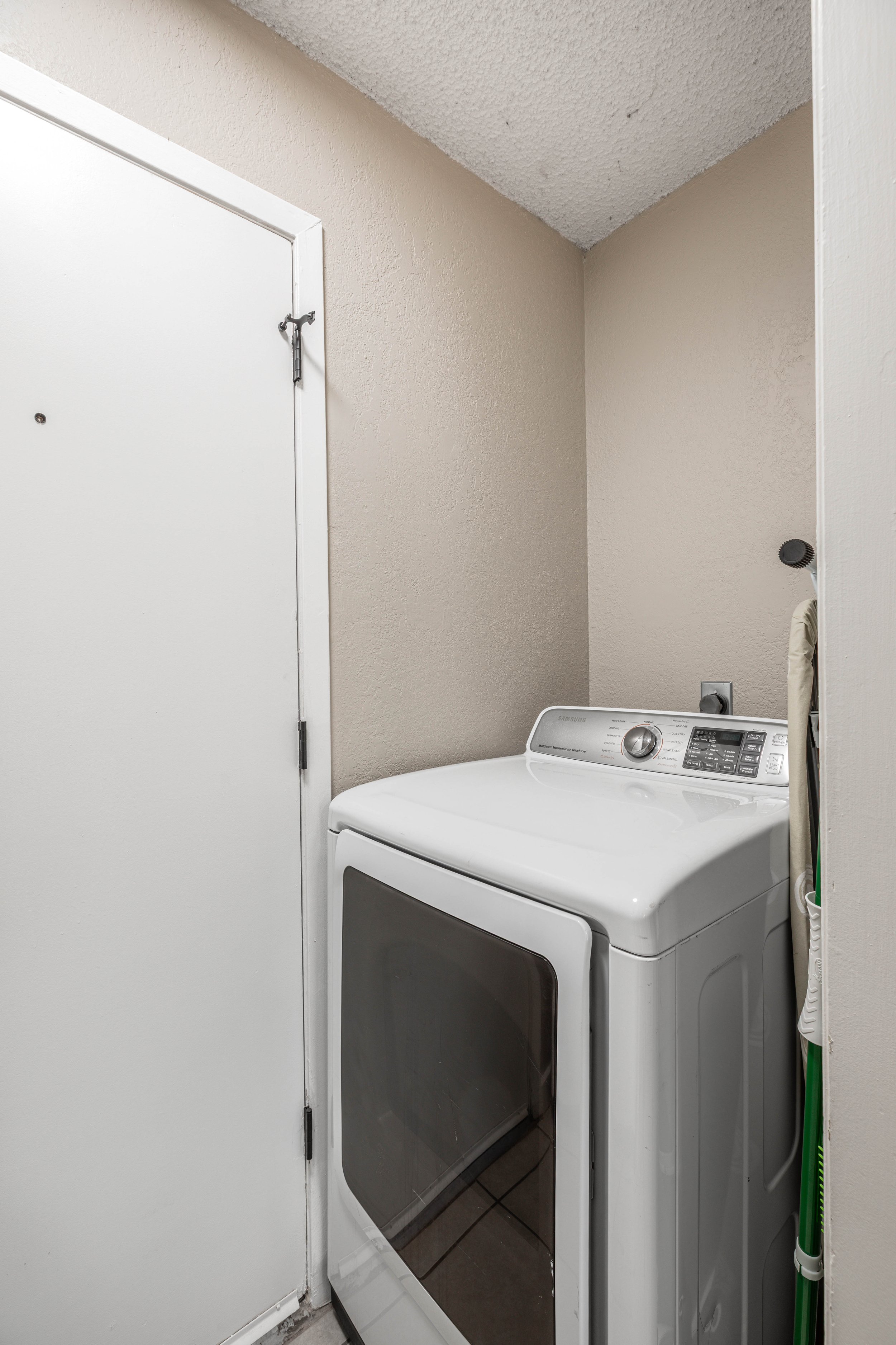 A laundry area with a white washing machine, a closed white door, beige walls, and a green broom leaning against the wall.