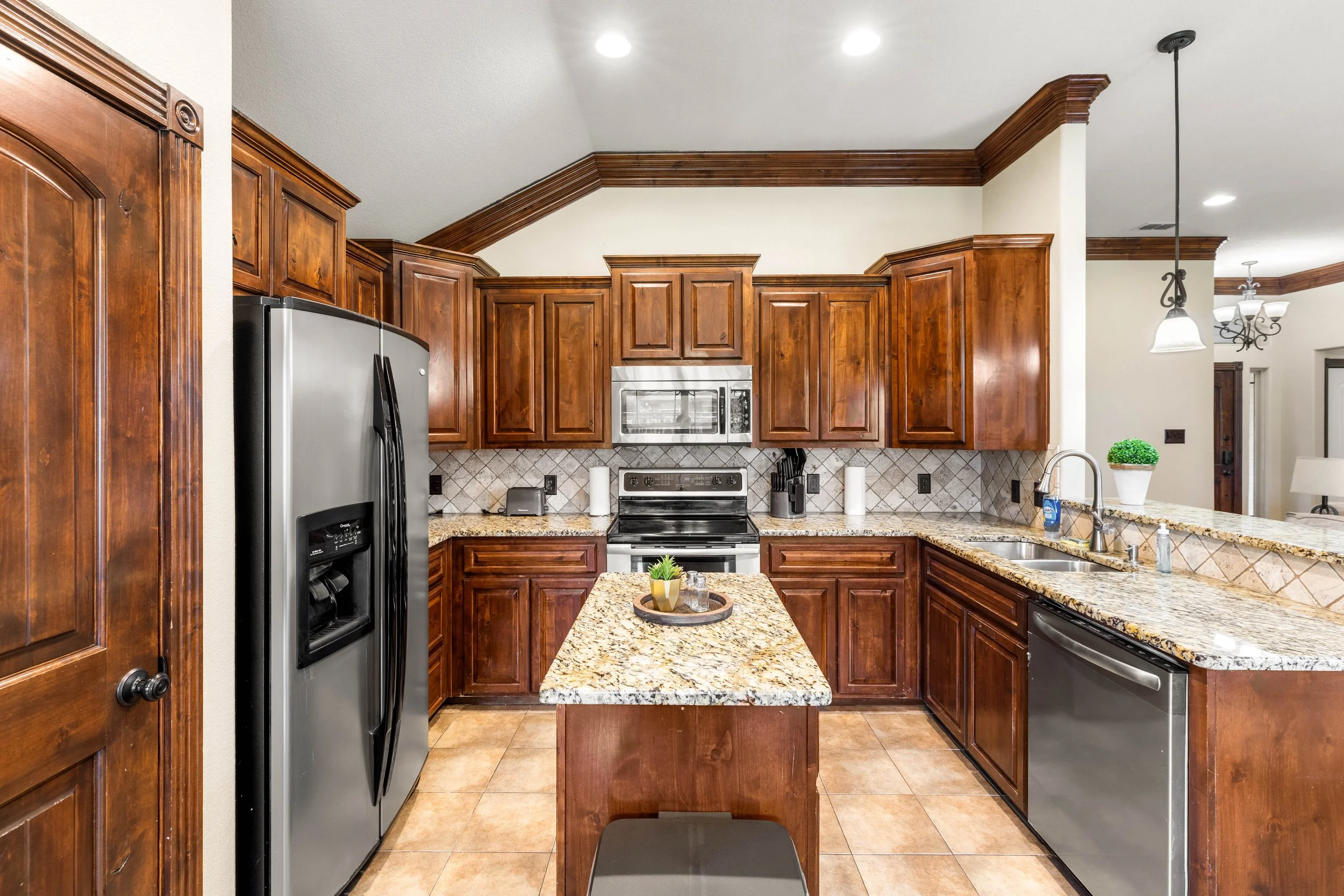 Kitchen with wooden cabinets, granite countertops, stainless steel appliances, a tiled backsplash, small potted plant on the island, and a hanging light fixture.