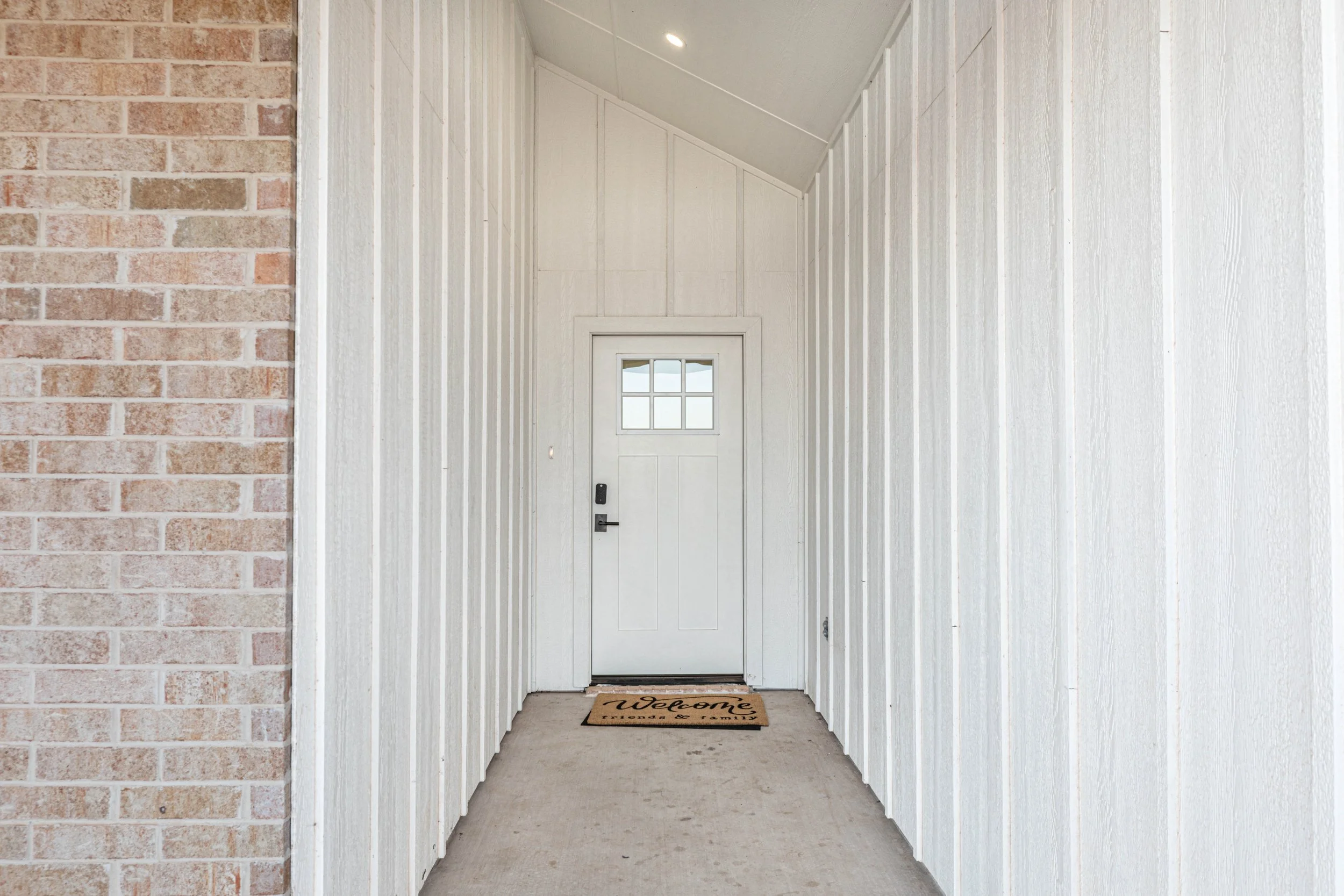 White front porch with a door, white paneling on the walls, and a welcome mat that reads 'welcome' in front of the door.