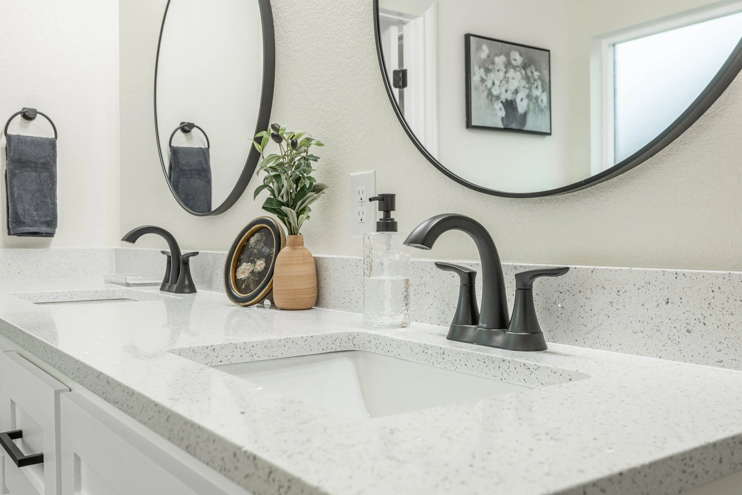 Bathroom vanity with white speckled countertop, black faucets, decorative vase with green leaves, a small framed picture, and a clear soap dispenser. Two round mirrors above the sinks, a dark gray towel on a ring, electrical outlets, a window, and a 